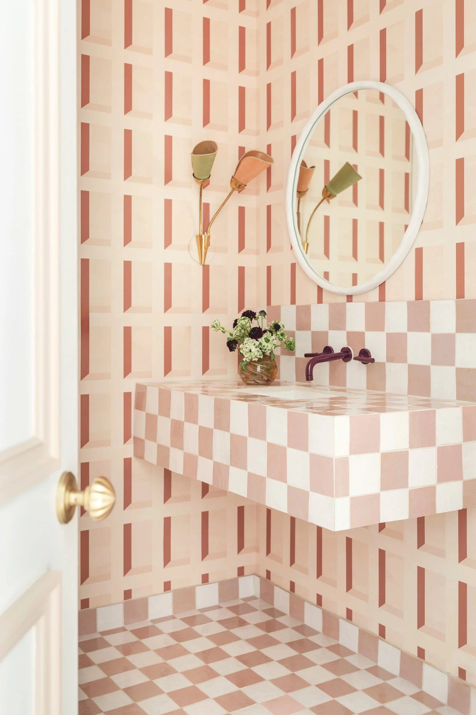 A powder room features a pink and white checkerboard tiled floating vanity, geometric 3D pattern wallpaper, a round white-framed mirror, and brass sconces.