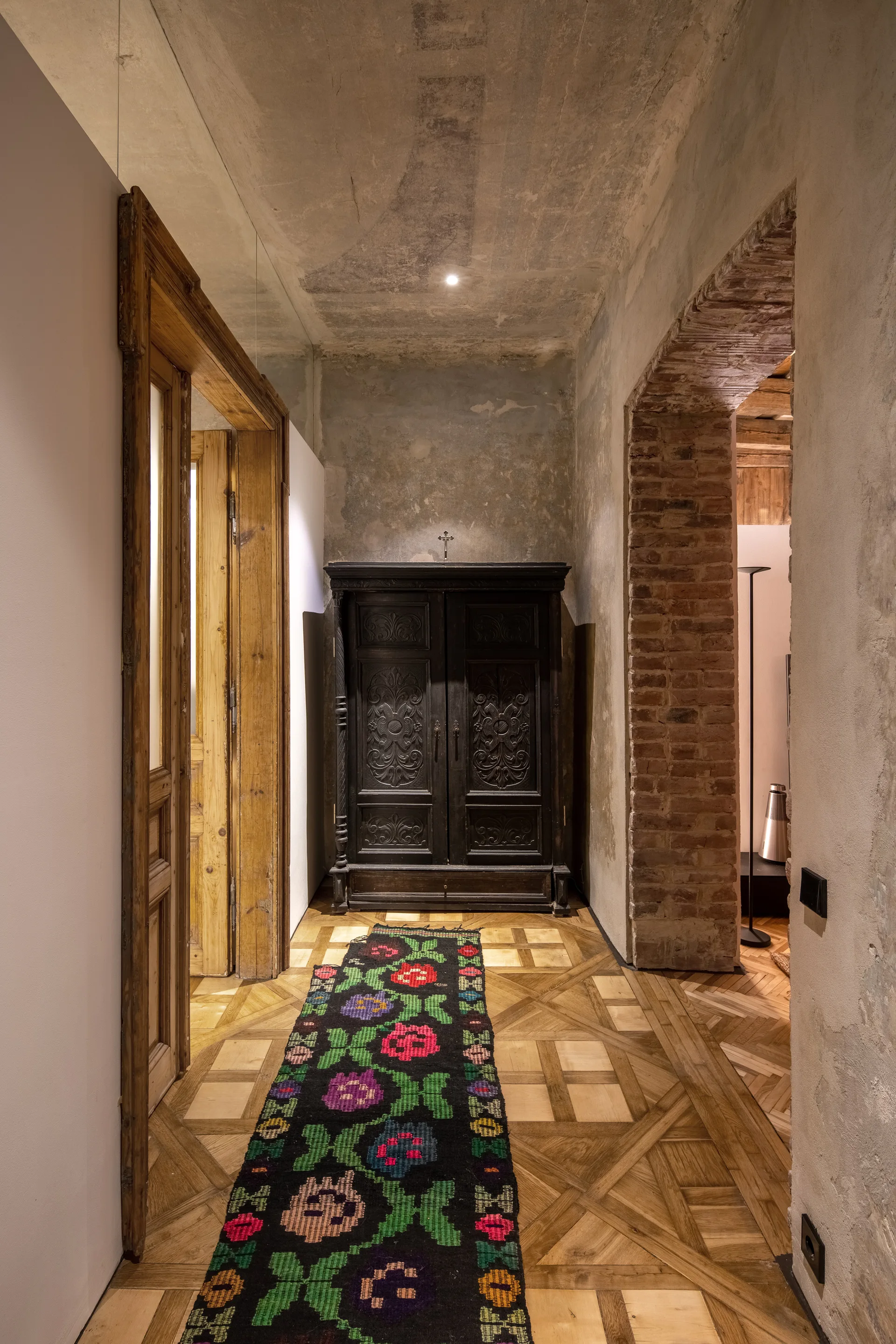 A rustic hallway features geometric parquet wood flooring, a colorful floral runner rug, a dark carved wood armoire, and an exposed brick archway.