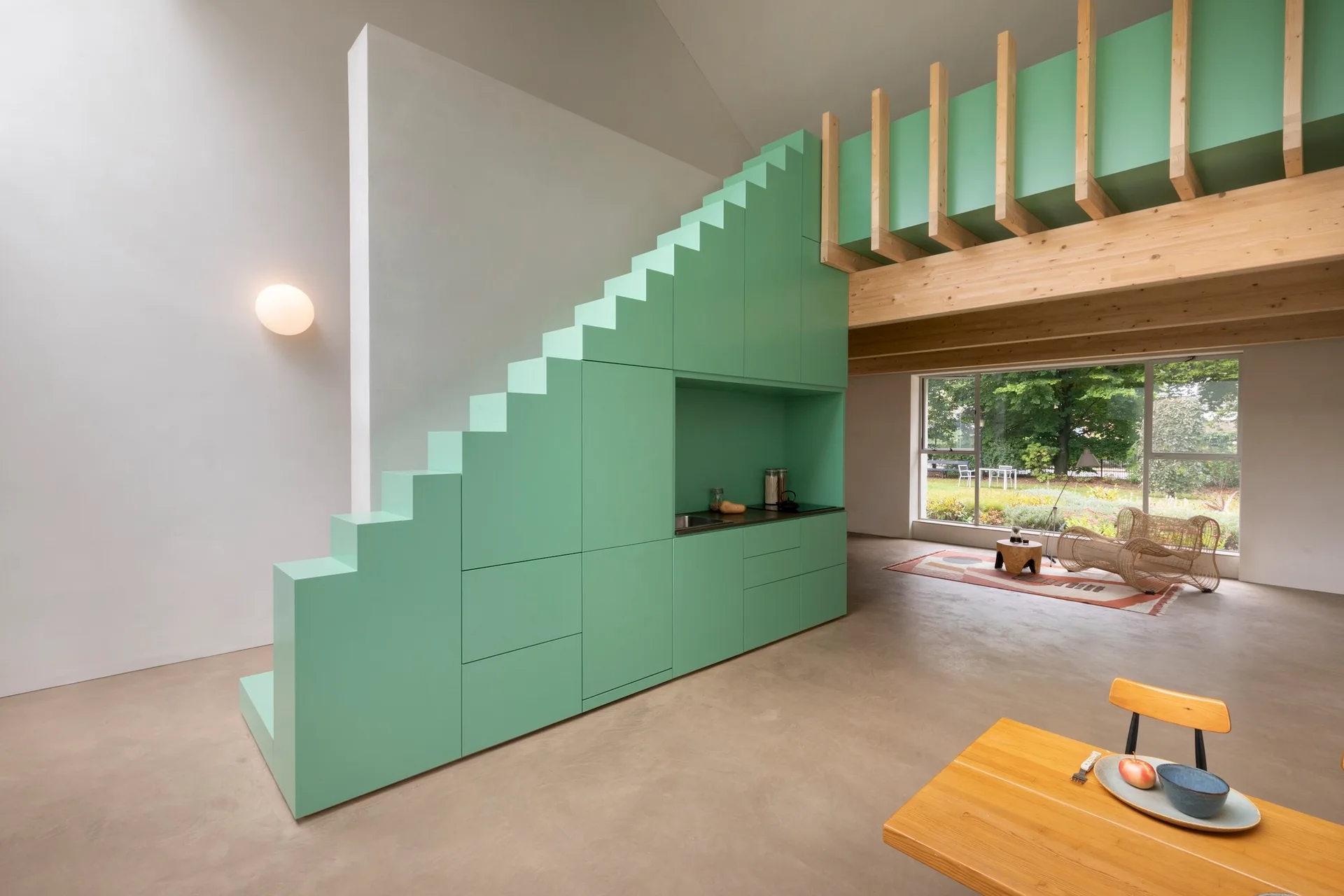 A mint green staircase featuring integrated storage and a small kitchen niche stands in an open-plan loft below a wooden mezzanine.