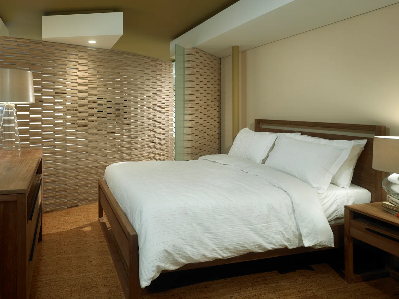 A modern bedroom features a wooden bed with white linens next to a decorative horizontal slatted wood divider and a cork-textured floor.