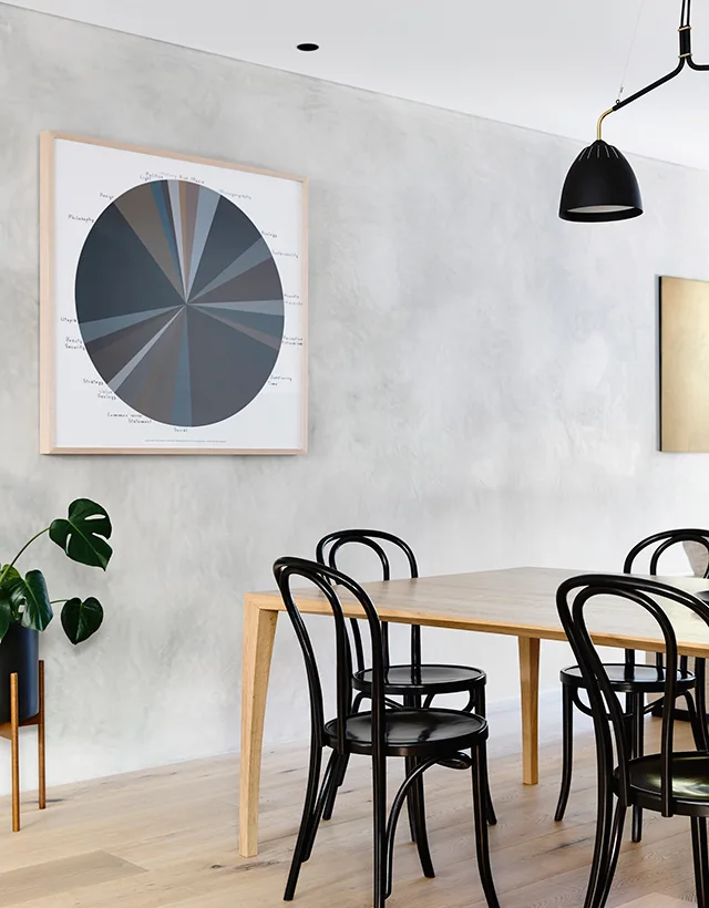 A minimalist dining room features a light wood table, black bentwood chairs, a large framed circular graphic print, and a textured grey plaster wall.
