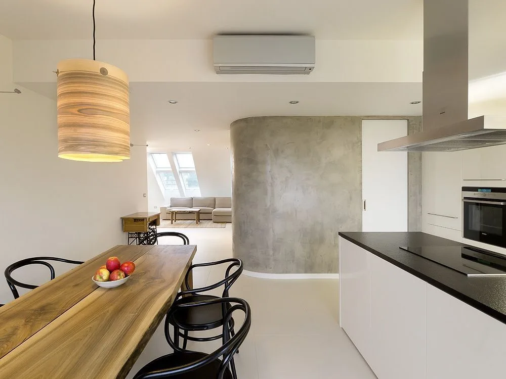 A modern open-concept kitchen and dining area features a live-edge wooden table, curved black chairs, and a smooth grey curved partition wall.