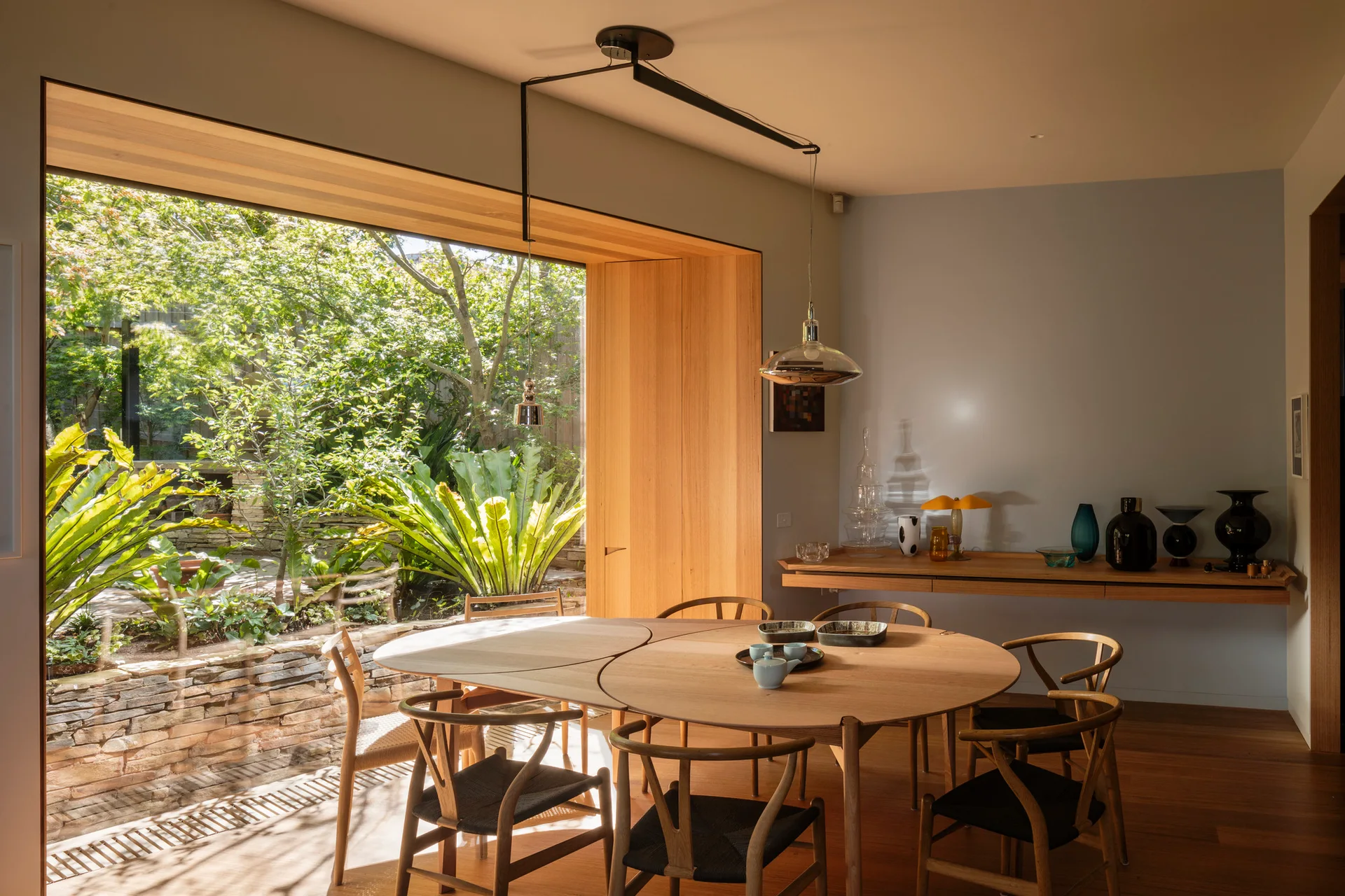 A round wooden dining table and chairs with black seats stand near a large wood-trimmed window overlooking a lush garden.