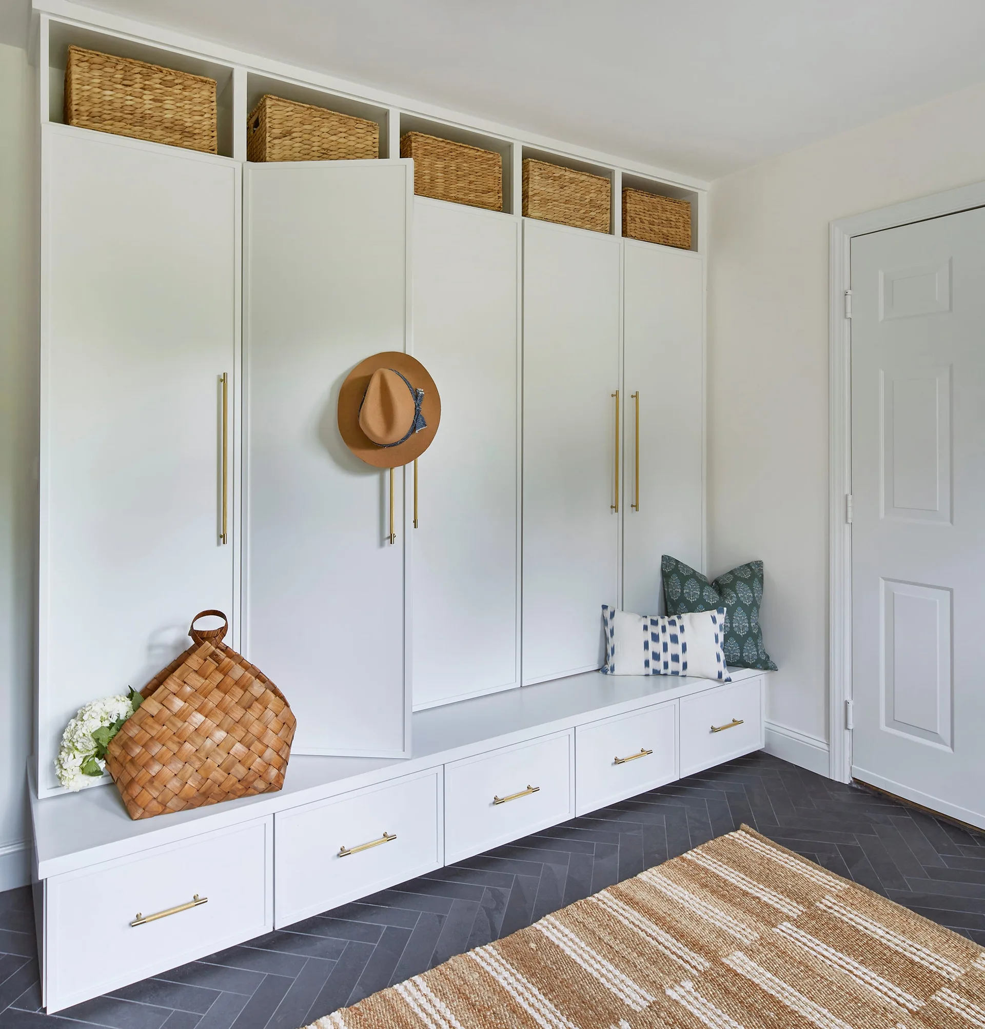 A white mudroom featuring custom built-in cabinets with an integrated bench, brass hardware, woven baskets, and dark grey herringbone floor tile.