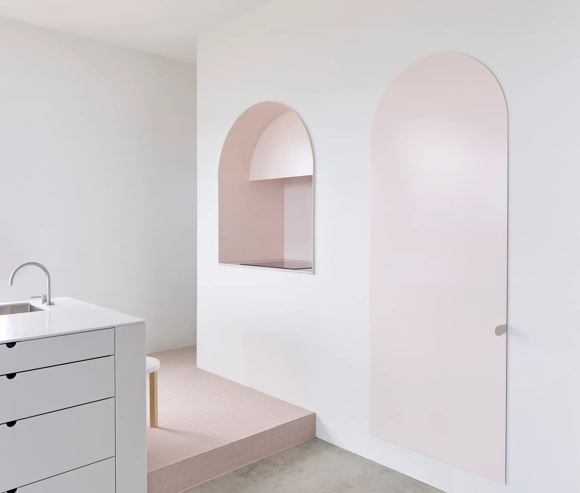 A minimalist kitchen featuring a white island, a pink arched flush door, and a matching arched alcove with a stovetop on a tiled platform.