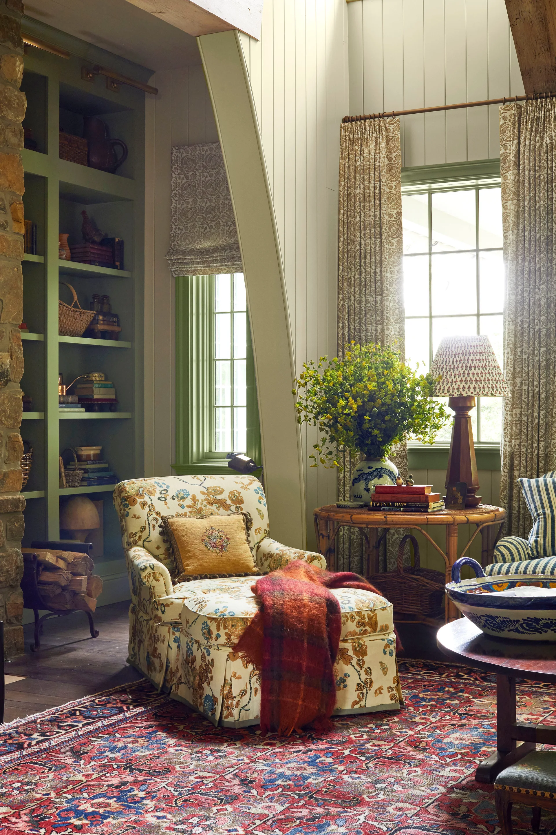 A cozy living space features a floral armchair with a matching ottoman, sage green built-in shelving, and a patterned area rug.