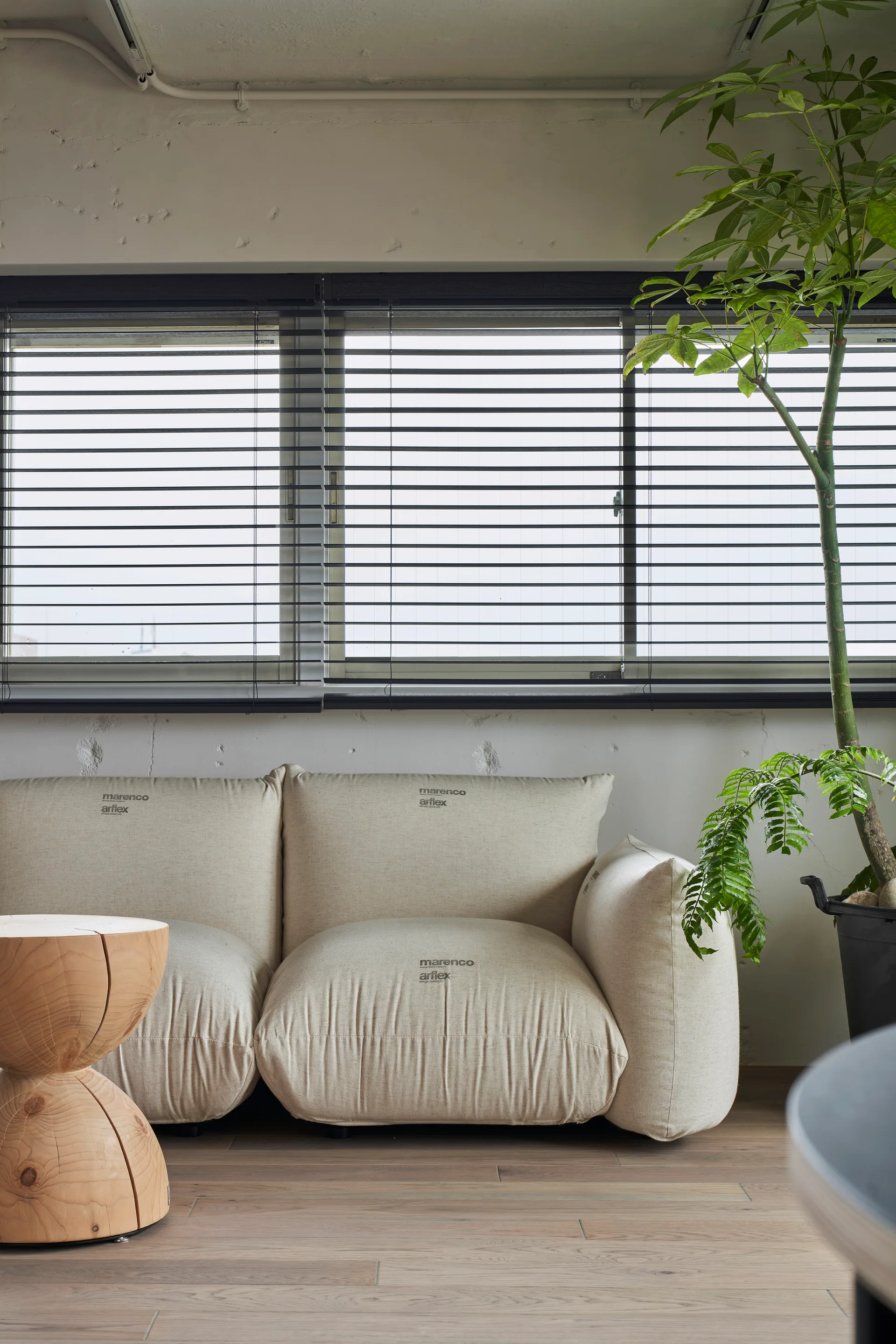A cream modular sofa sits on light wood floors beside a sculptural wooden side table and a large window with black venetian blinds.