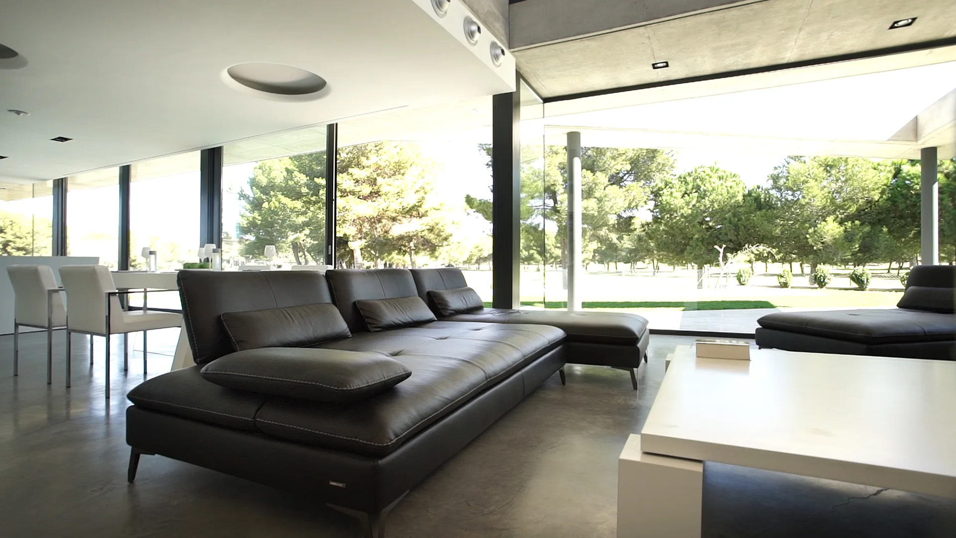 A modern living room features a low black leather sectional sofa, a white coffee table, and large glass walls overlooking green trees.