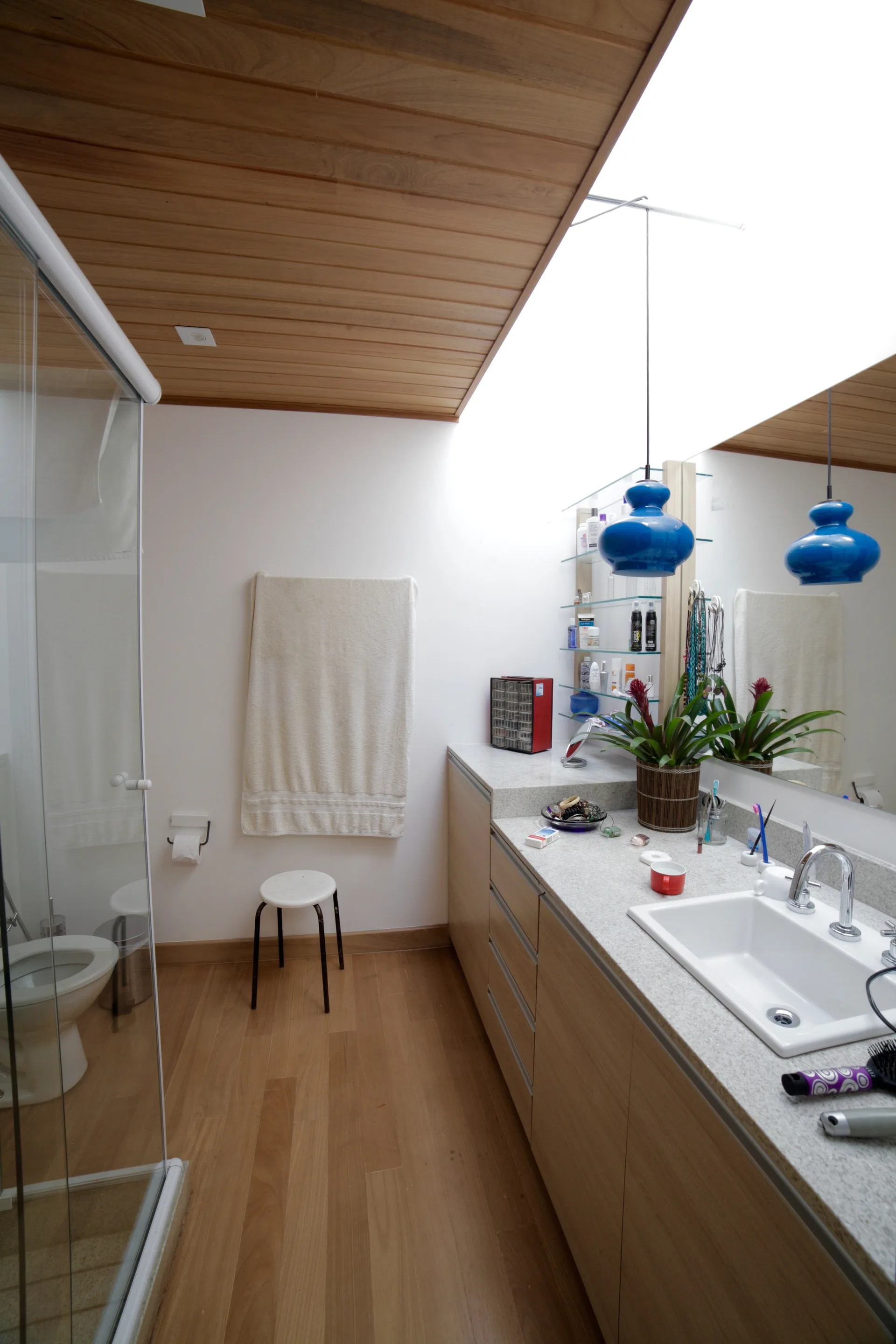 A modern bathroom features a wood plank ceiling, a long wood vanity under a bright skylight, and blue glass pendant lights hanging near a mirror.