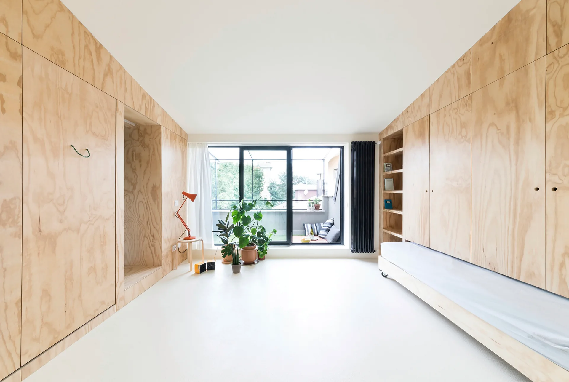 A minimalist bedroom featuring light wood plywood cabinets cladding opposing walls, a built-in daybed, and a large glass door leading to a balcony.