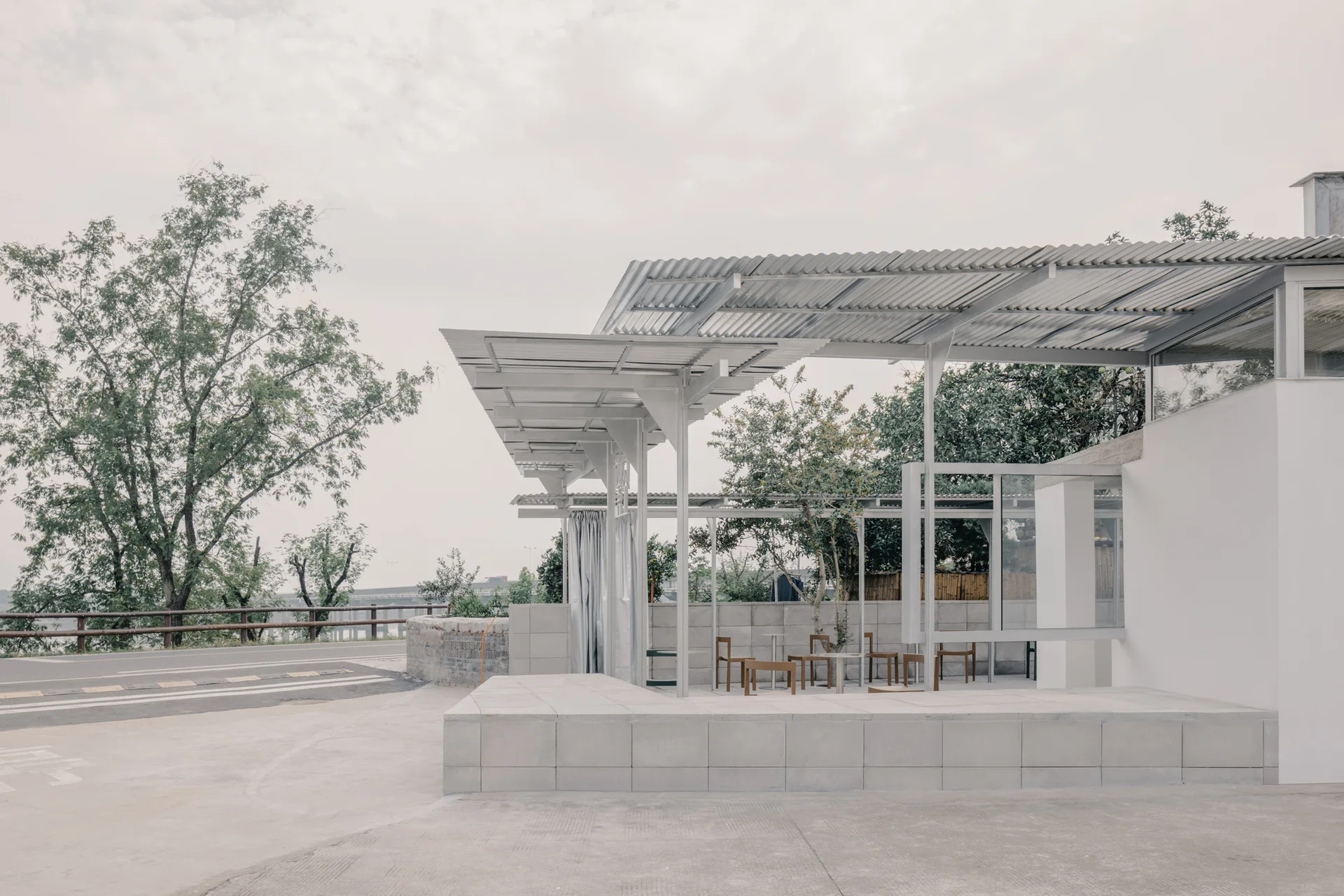 A minimalist outdoor patio with a corrugated metal canopy, concrete block foundation, and simple wood seating under an overcast sky.