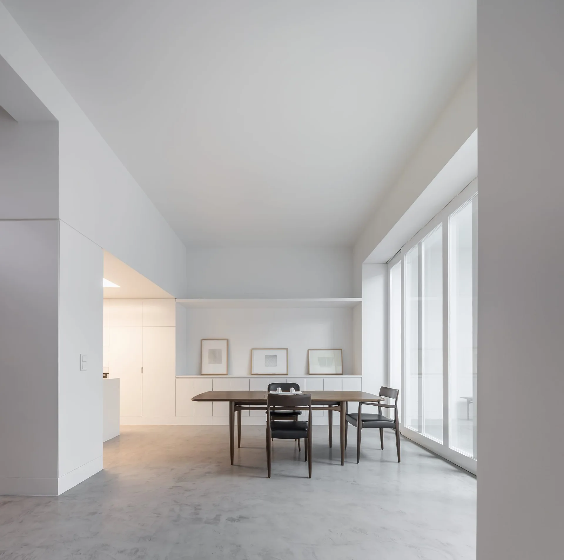 A minimalist dining room features a dark wood table and chairs on a grey concrete floor next to large glass windows.