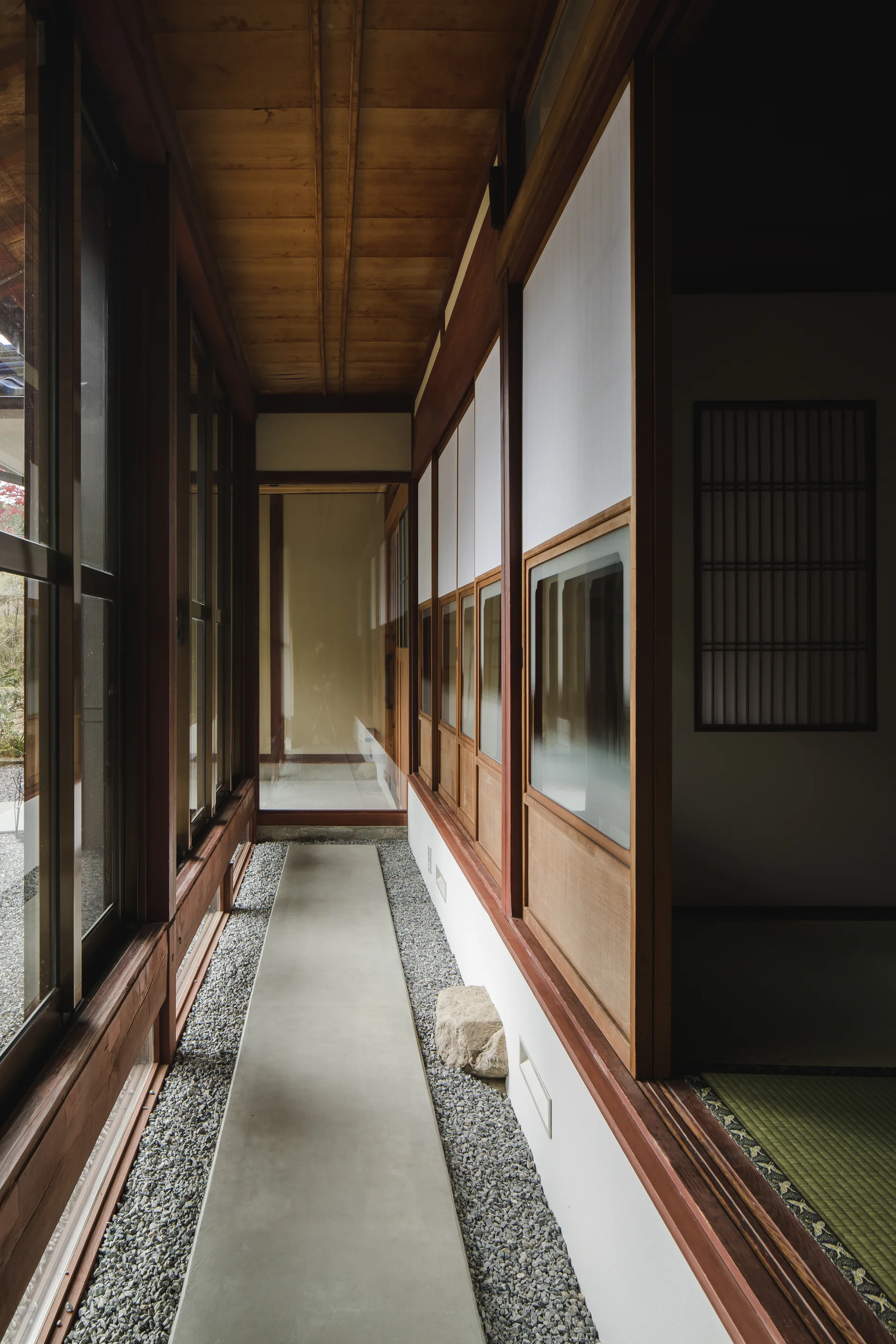 A narrow Japanese-style corridor featuring a central concrete pathway laid over grey gravel, flanked by sliding wooden doors and windows.