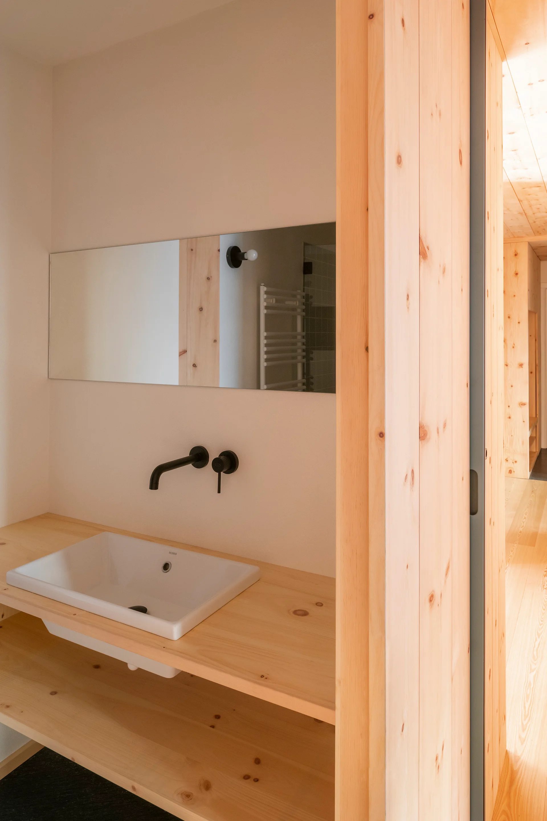 A minimalist bathroom vanity features a white ceramic sink on a natural pine wood counter with a wall-mounted matte black faucet and a frameless mirror.