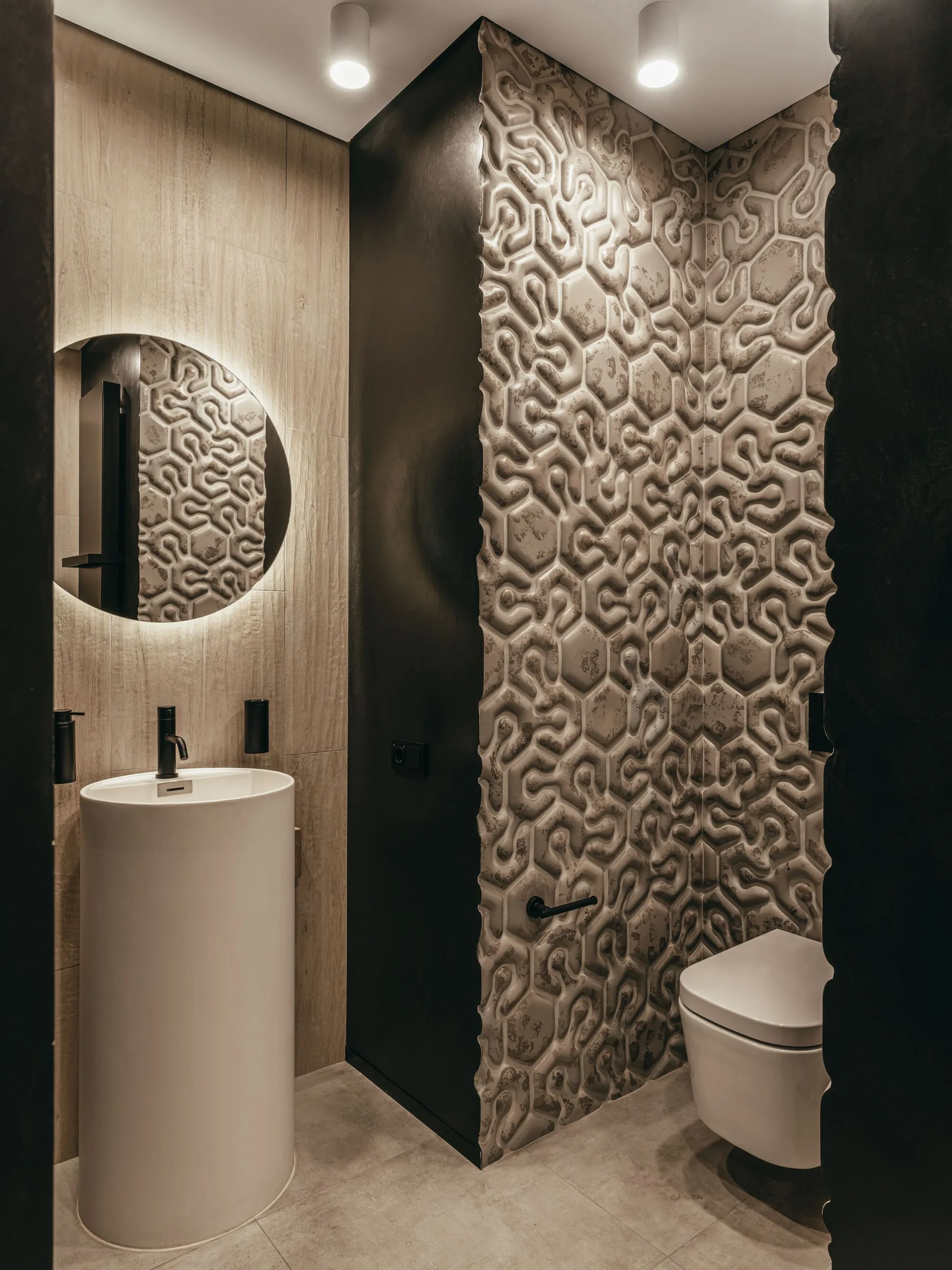 A modern powder room featuring a white cylindrical pedestal sink, a circular backlit mirror, and a wall of highly textured 3D geometric tiling.