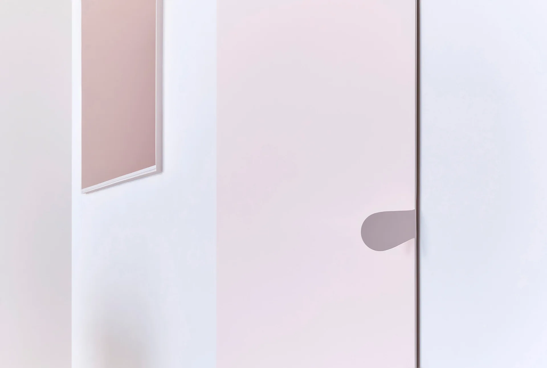 A close-up of a minimalist pale pink door with a unique organic-shaped handle next to a dusty rose rectangular frame on a white wall.