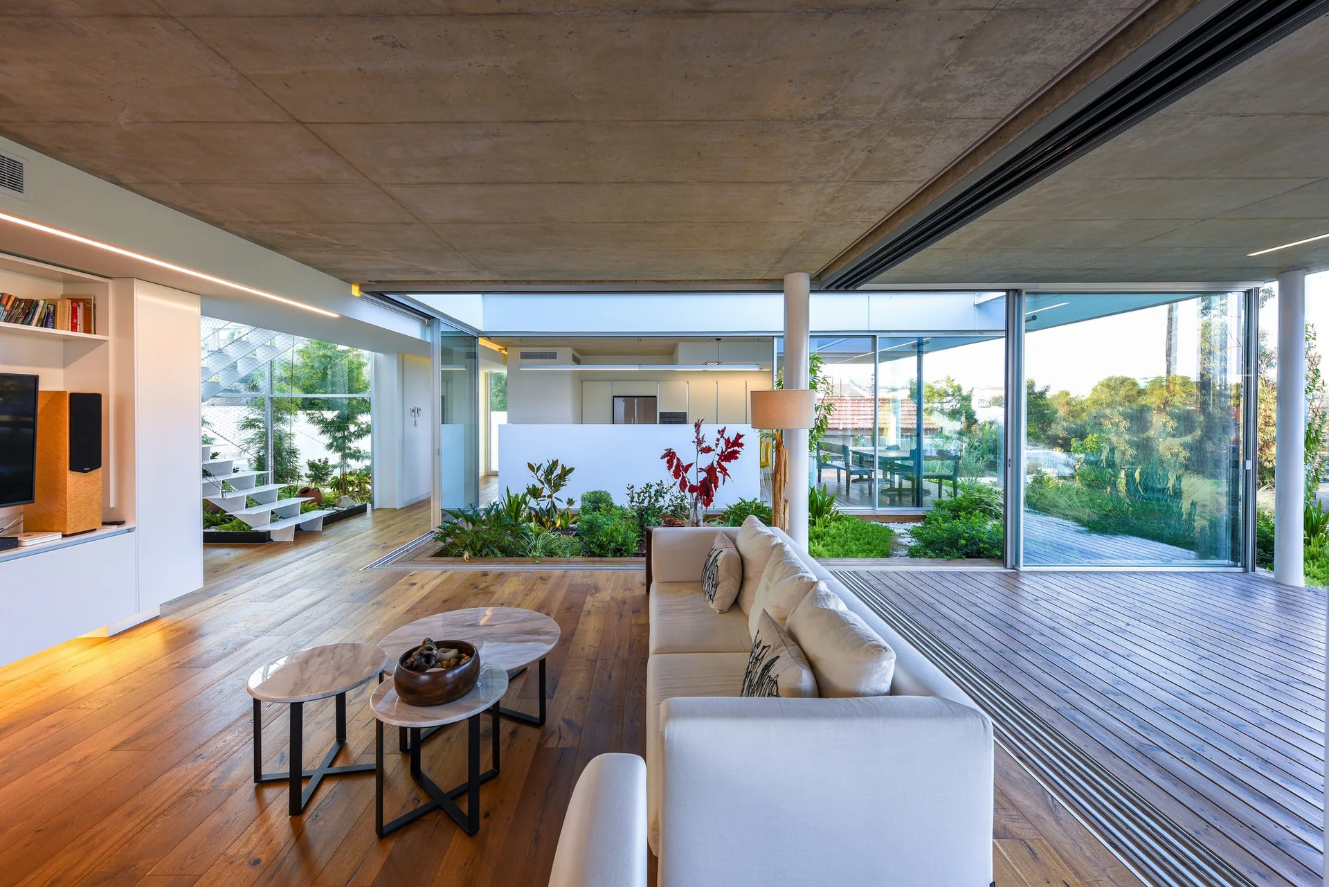 A modern living room features a cream sofa, nesting coffee tables, a raw concrete ceiling, and sliding glass doors opening to a central courtyard garden.