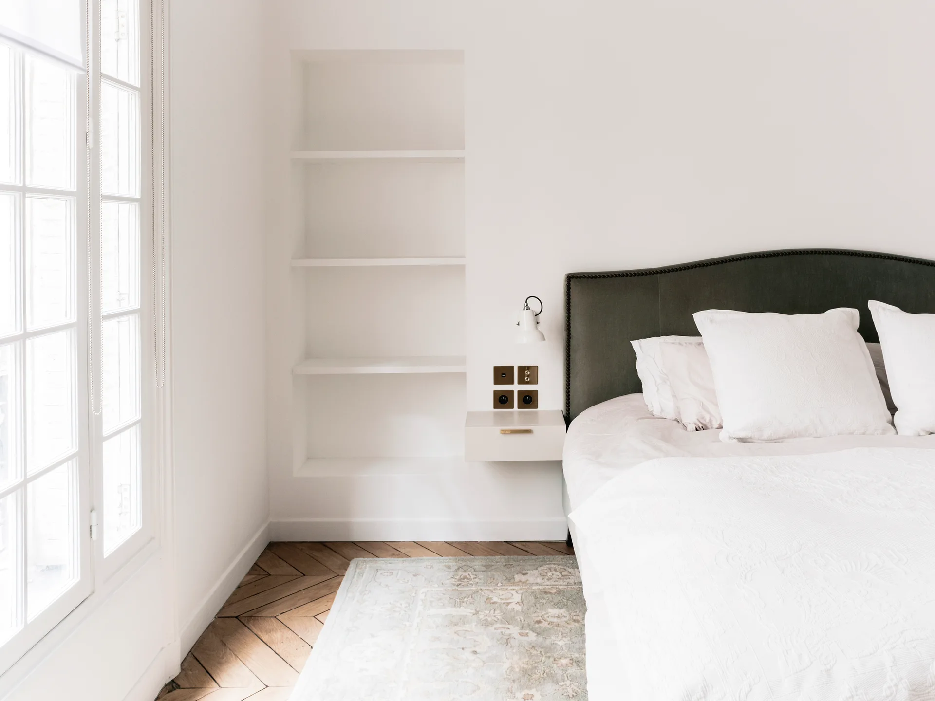 A bright bedroom featuring a dark olive headboard, a white wall-mounted nightstand below a recessed shelving niche, and herringbone wood floors.