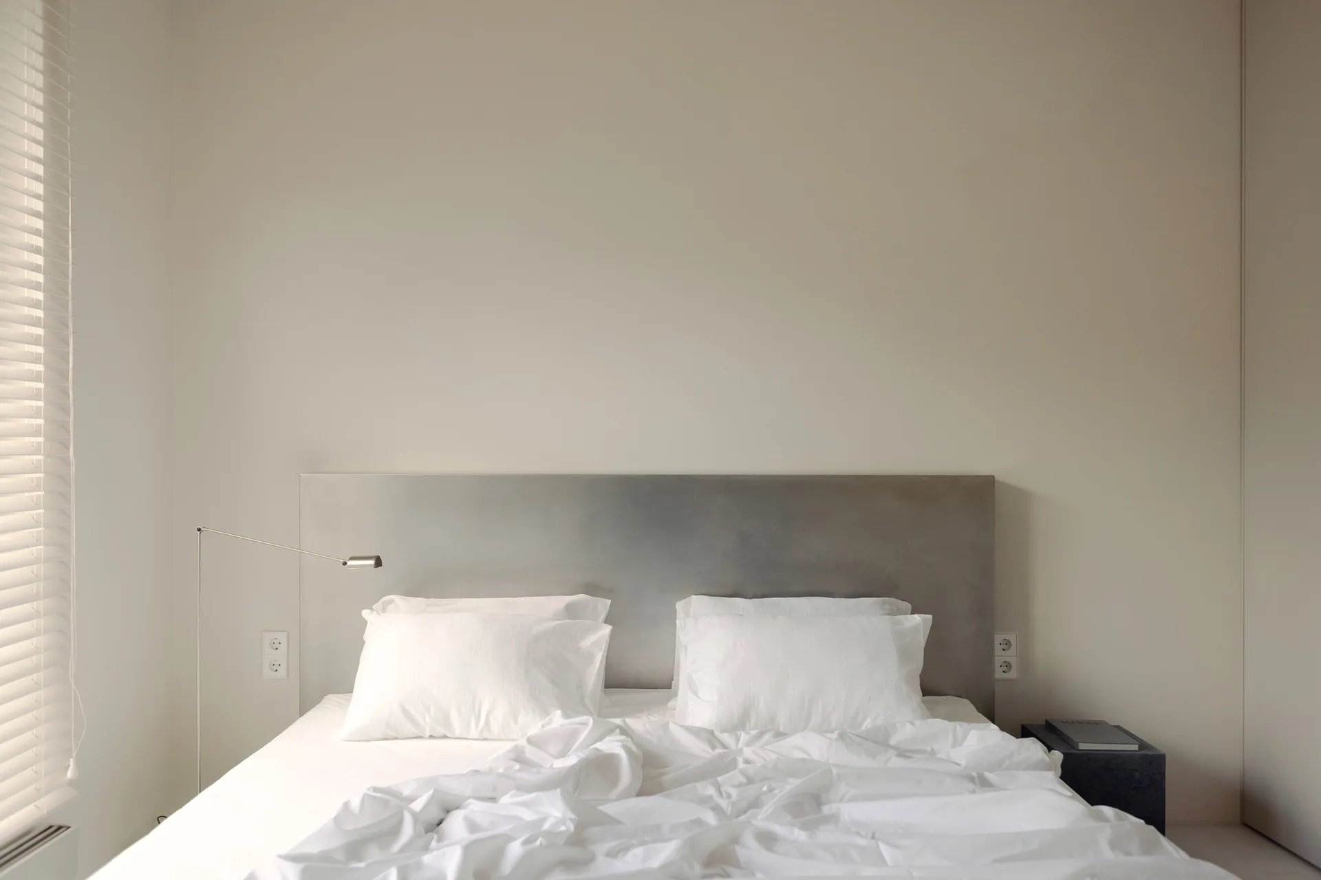 A minimalist bed with white linens and a wide grey metal headboard stands against an off-white wall near venetian window blinds.