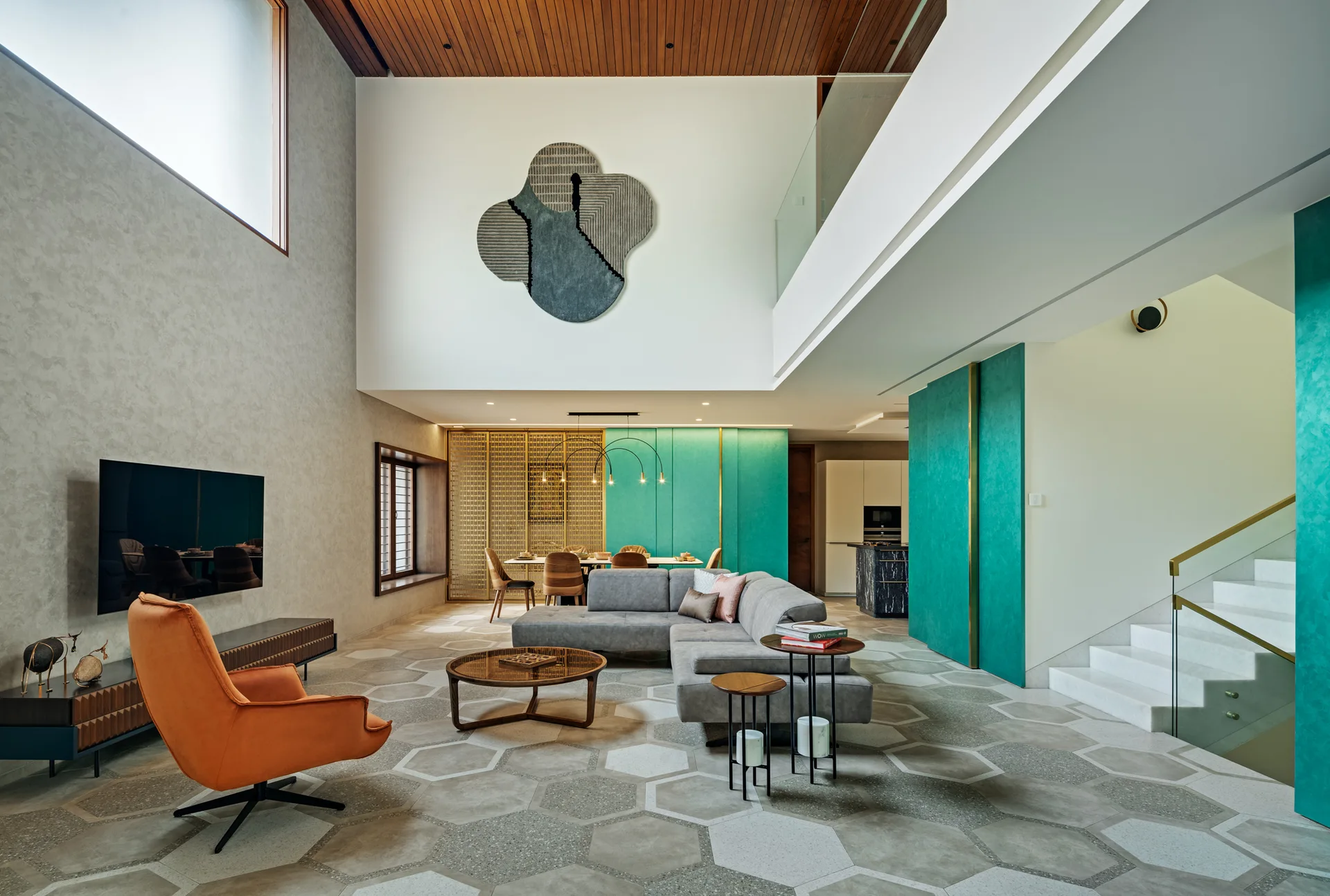 A modern double-height living room featuring a grey sectional, an orange swivel chair, hexagonal floor tiles, and striking teal accent panels.