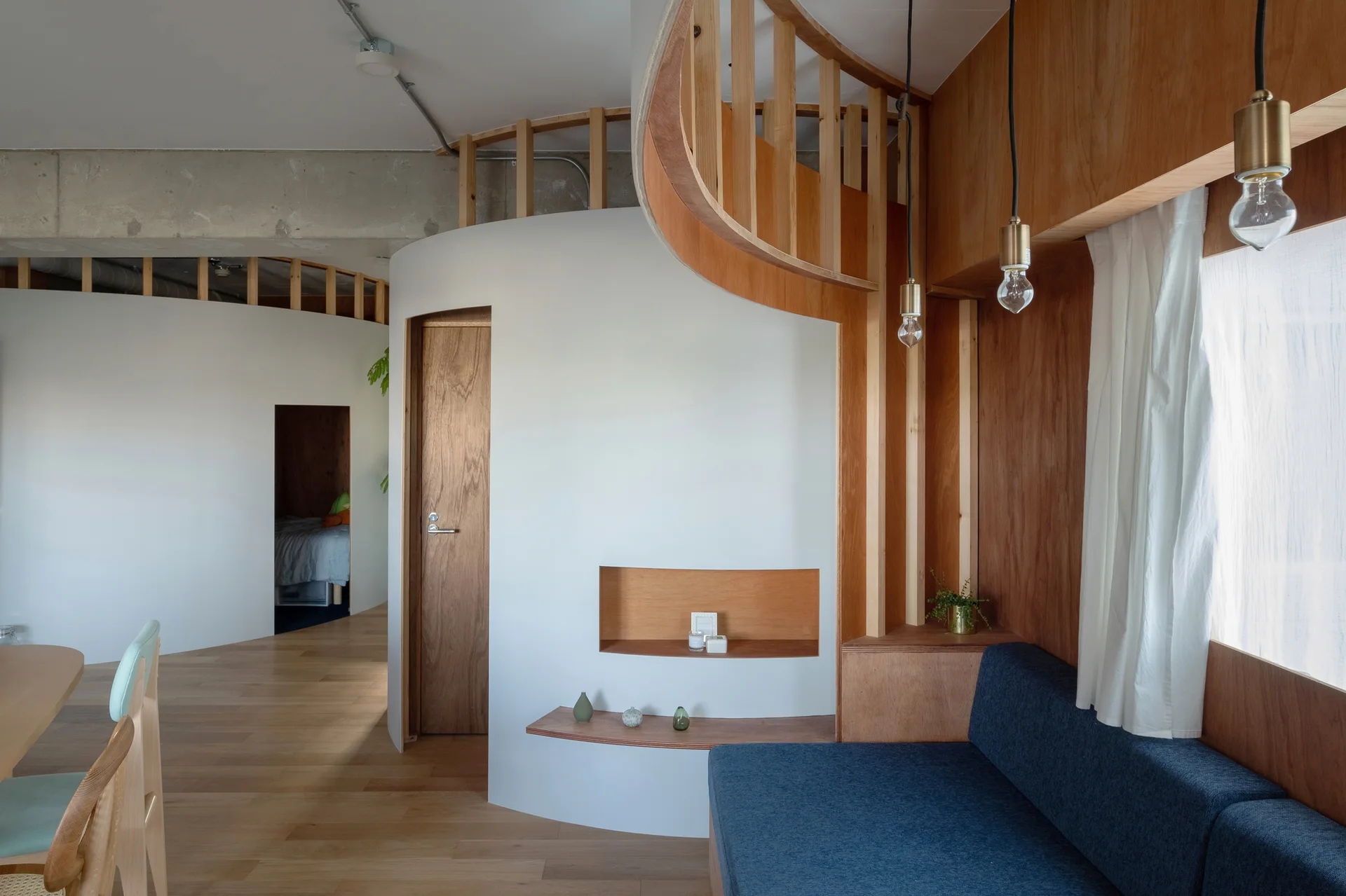A modern living space features a curved white partition wall, built-in navy blue sofa, recessed wooden shelving, and light wood flooring.
