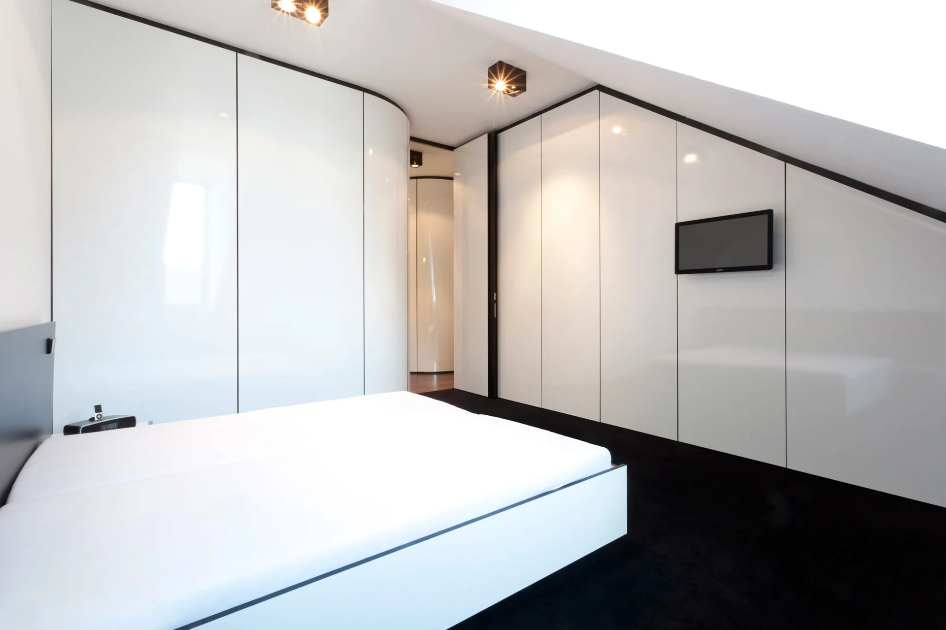 A minimalist attic bedroom featuring a white platform bed, high-gloss white built-in wardrobes, a slanted ceiling, and a black carpet.