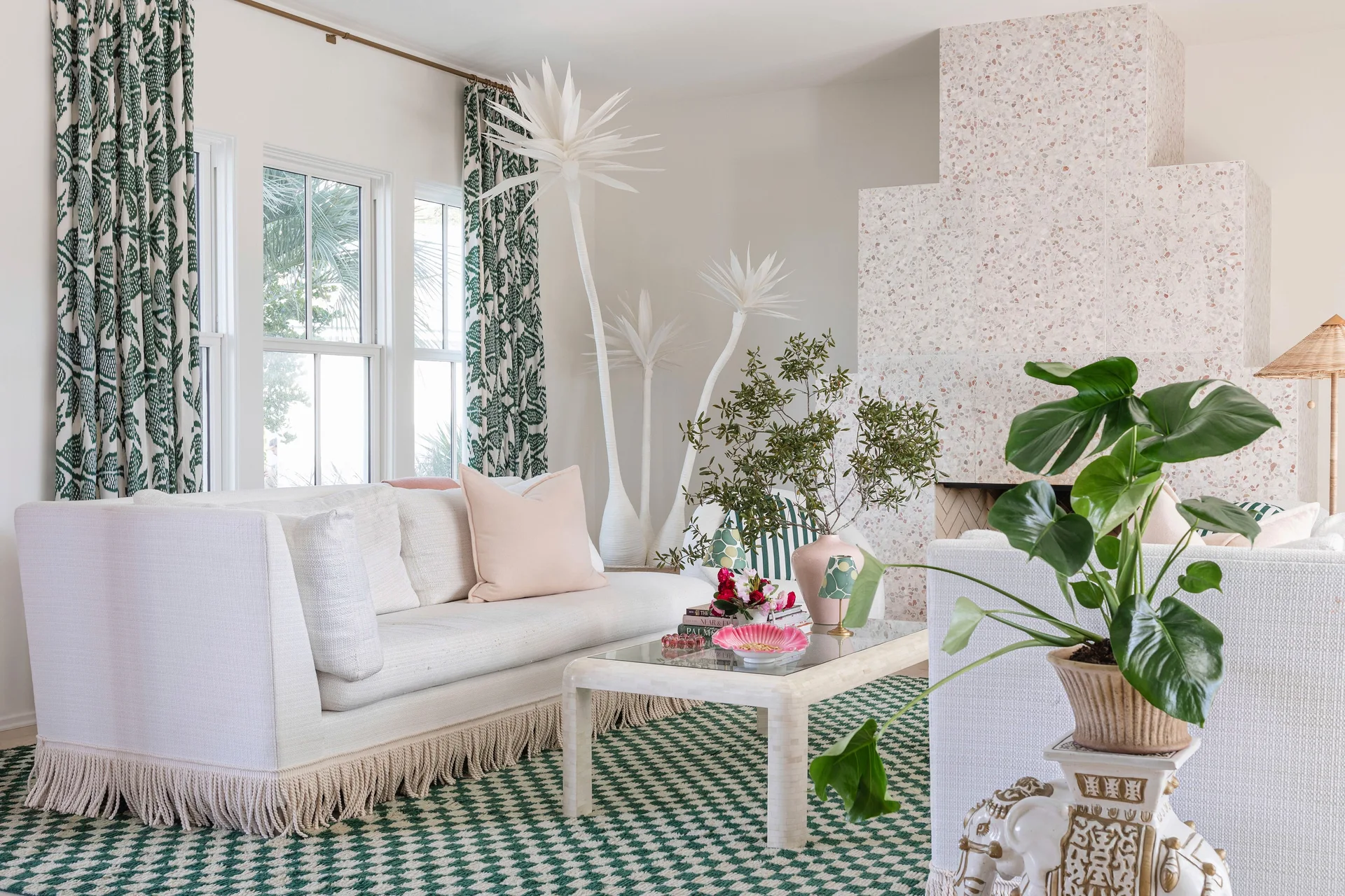 A bright living room features a white fringed sofa, a green and white houndstooth rug, and a tall speckled pink terrazzo fireplace surround.