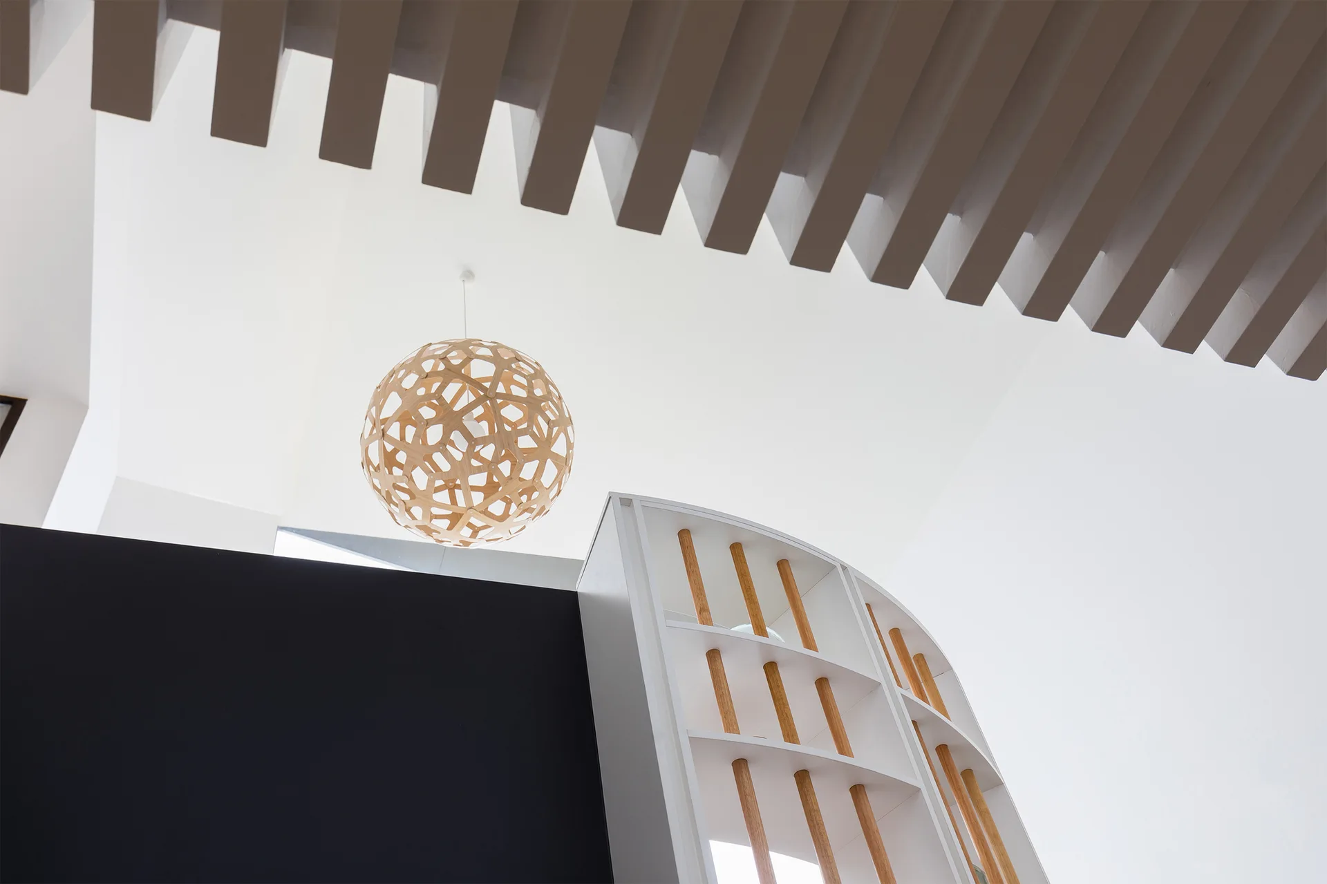 Low angle view of a spherical geometric wooden pendant light hanging near a slatted ceiling and a curved white shelving unit with wooden dowels.
