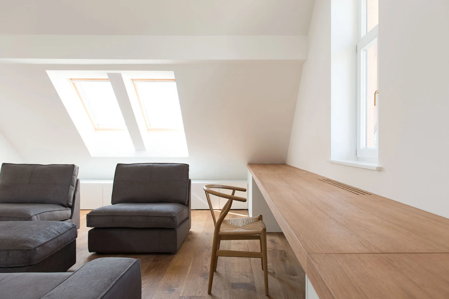 An attic living space with slanted ceilings, skylights, dark gray upholstered lounge chairs, and a long built-in light wood desk with a wooden chair.