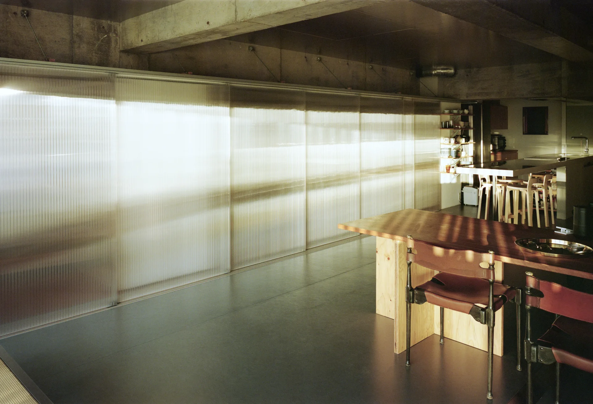 A modern dining space features a wooden table with leather chairs alongside a large translucent ribbed sliding partition and exposed concrete ceiling.