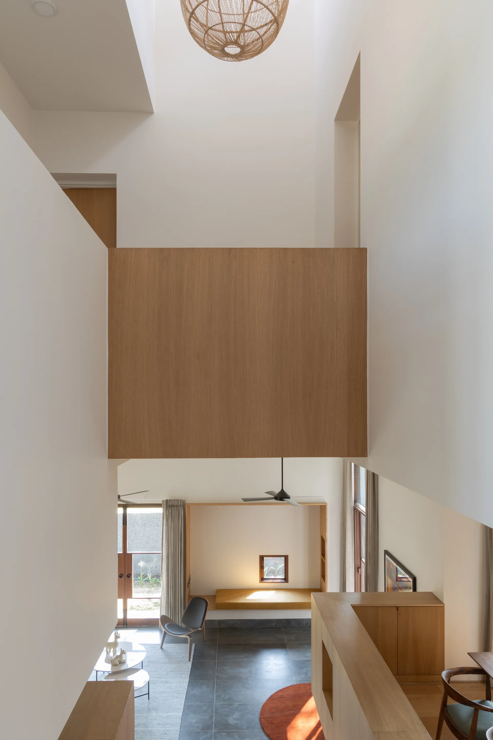 High-angle view of a modern double-height living room featuring a central wood-paneled landing bridge, white walls, and a spherical wicker pendant light.