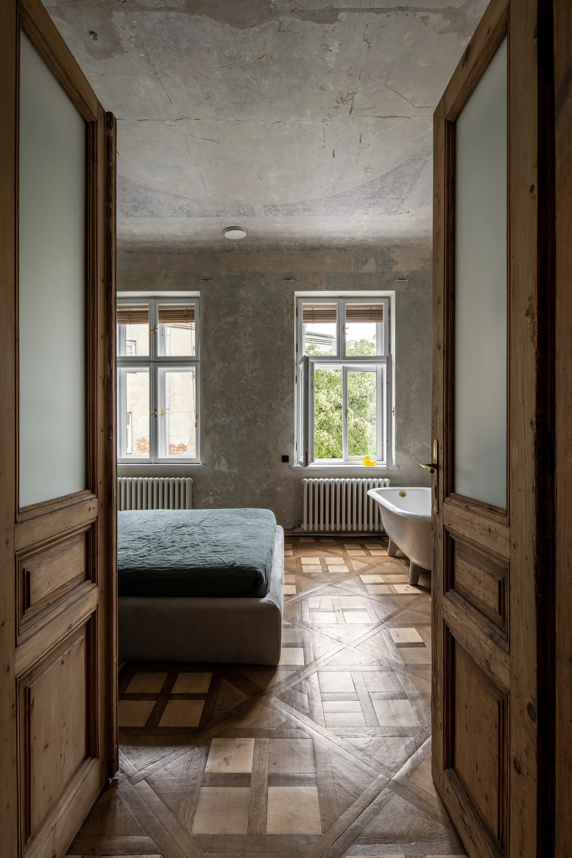 A view through wooden double doors into a bedroom featuring a bed, a white clawfoot bathtub, distressed grey walls, and patterned parquet flooring.