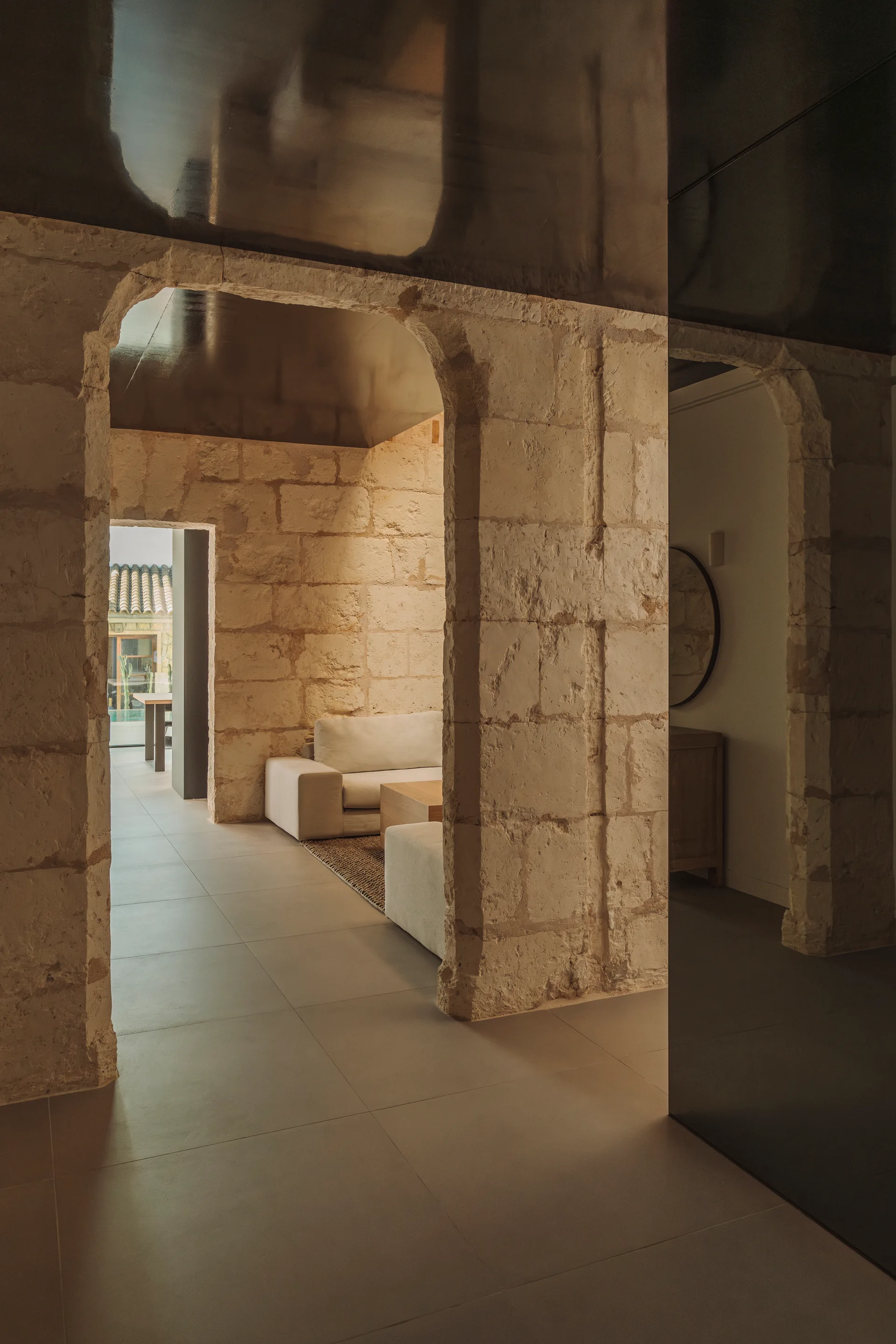 A residential interior features textured tan stone archways, a dark reflective ceiling, a light cream sofa, and beige tiled floors.