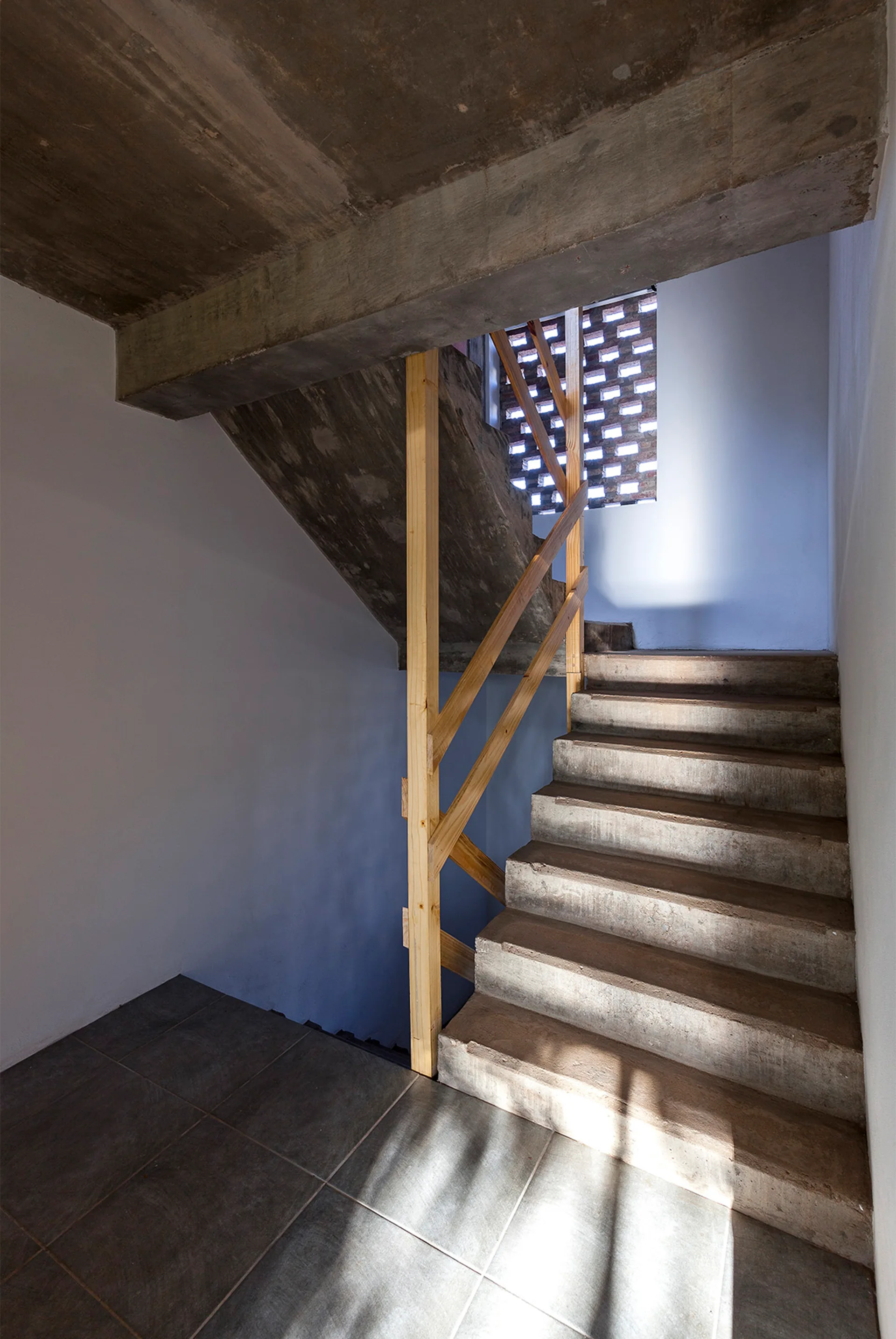 A raw concrete staircase with a simple light wood railing sits next to white walls and a perforated brick window screen that filters natural light.
