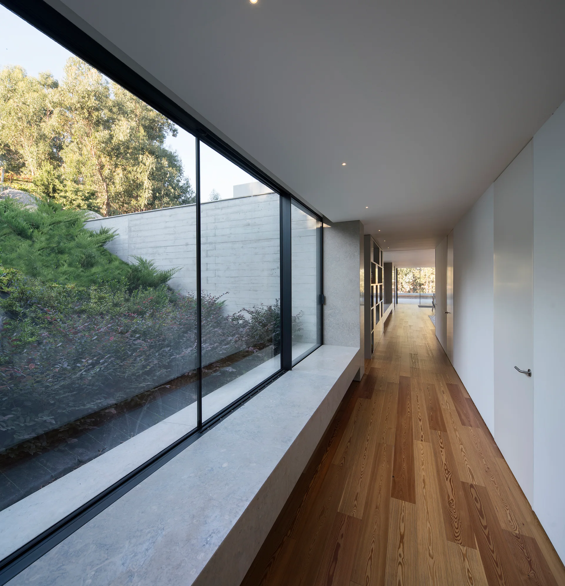 A minimalist residential hallway features warm wood plank flooring, flush white doors, and a long floor-to-ceiling glass wall showing an outdoor garden.