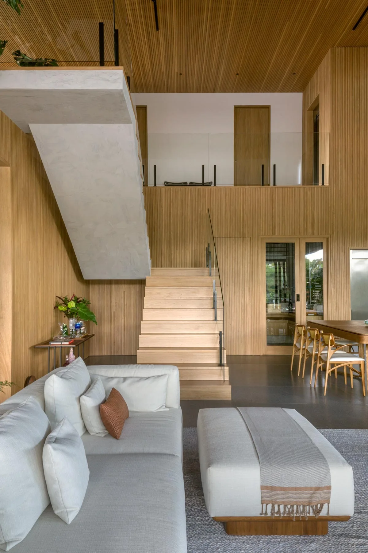 A contemporary living room featuring a white sofa, a matching ottoman, a wooden staircase with glass railings, and vertical wood-slatted walls.
