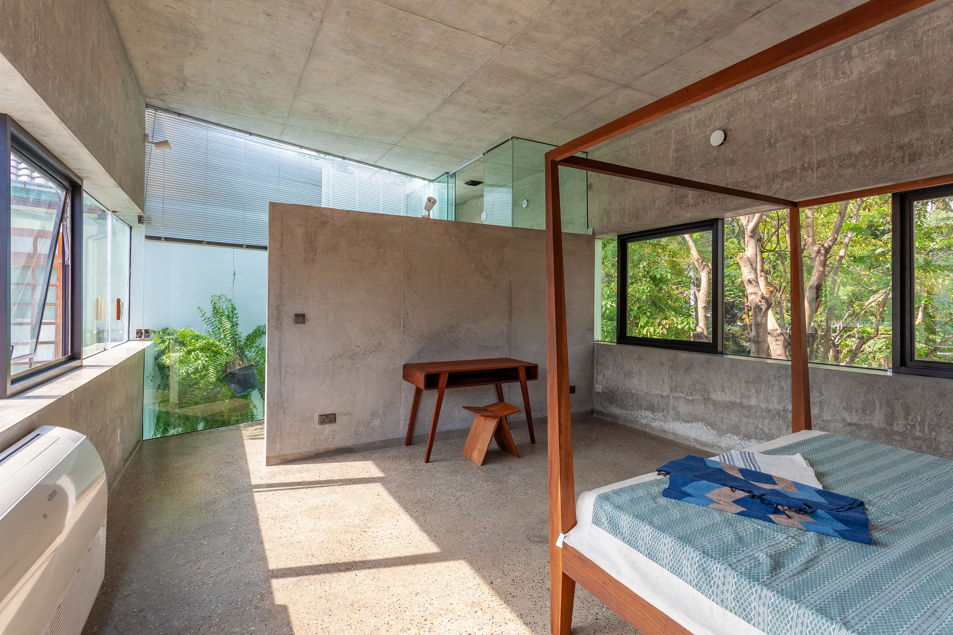 A minimalist bedroom featuring raw concrete walls, a wooden four-poster bed, a small desk with a stool, and large windows facing green trees.
