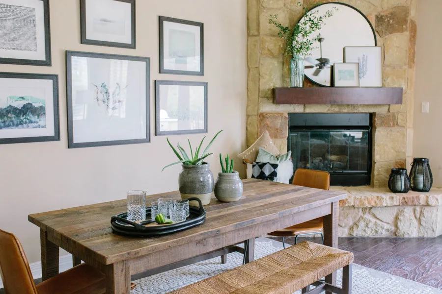A rustic wooden dining table with a woven bench and leather chairs is positioned near a stone fireplace and a gallery wall of framed art.