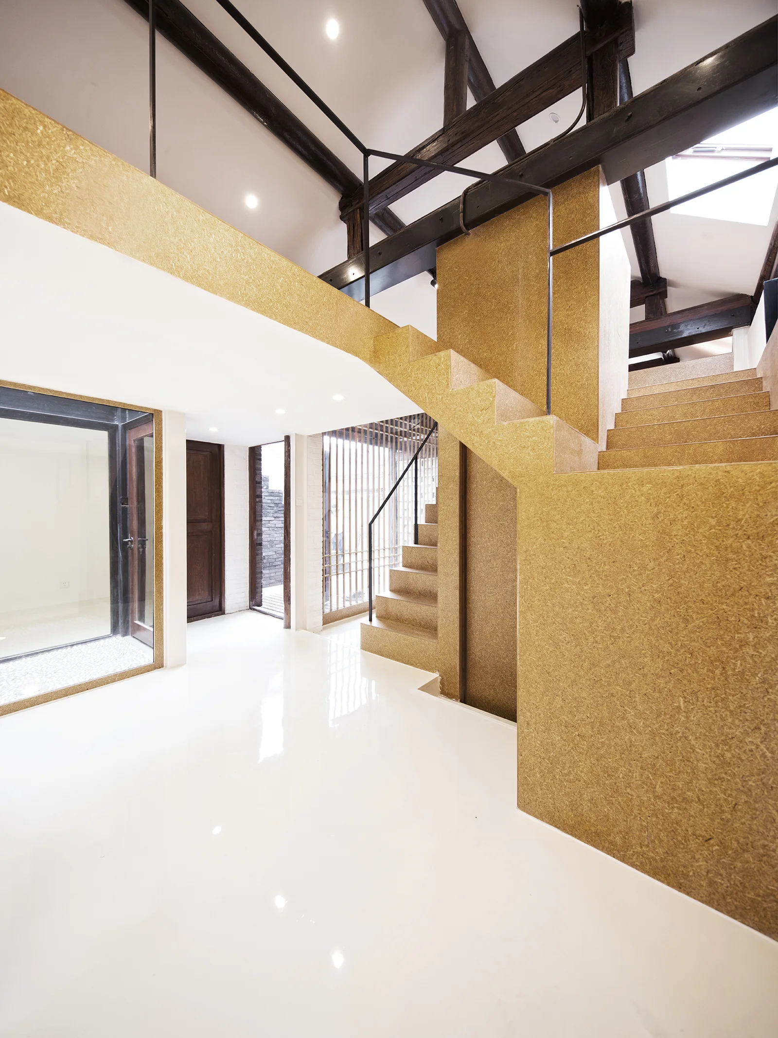 An open interior featuring a central staircase clad in textured composite board, a glossy white floor, and exposed dark timber ceiling beams.