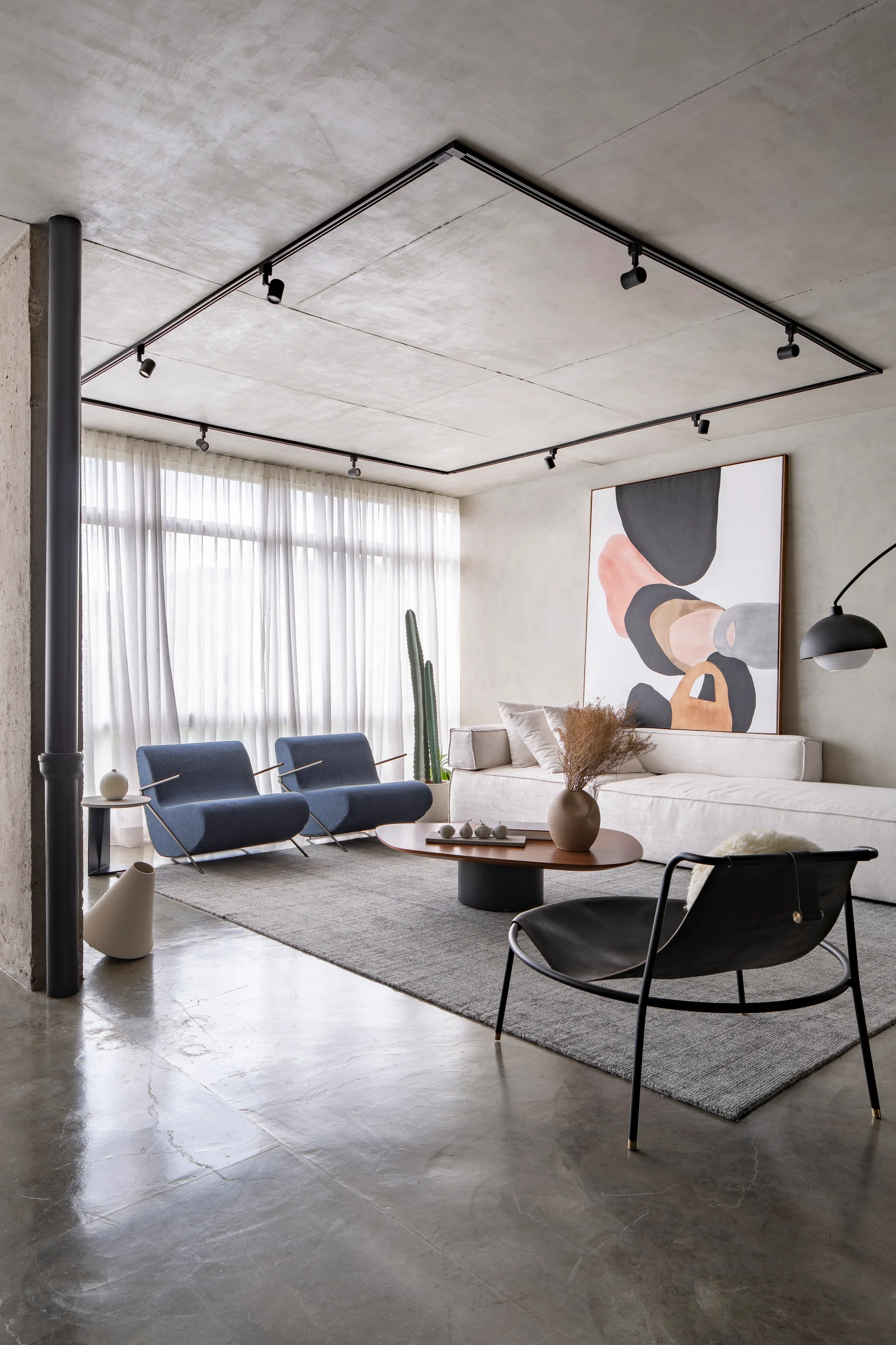 A modern living room features a white sofa, a pair of blue armchairs, and a low wood coffee table beneath a concrete ceiling with black track lighting.