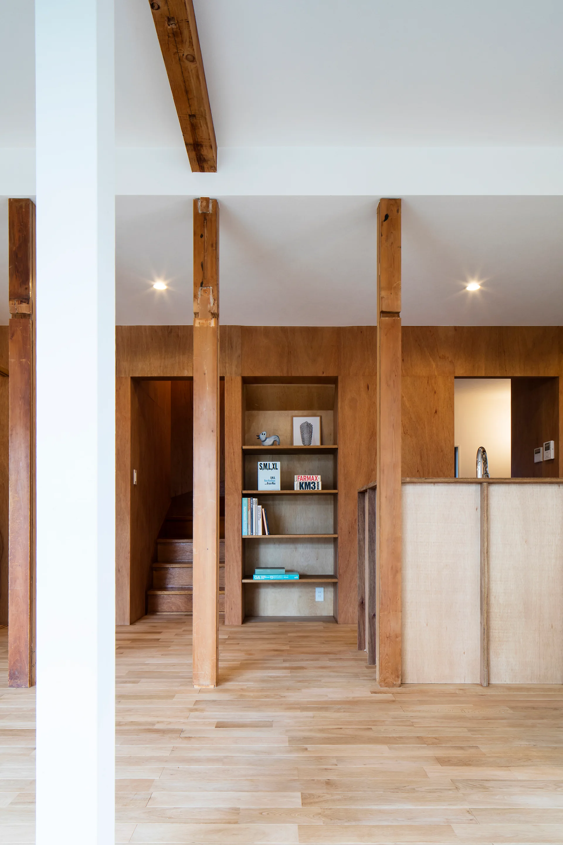 An interior space features light plywood paneled walls, a built-in recessed wooden bookshelf, exposed timber structural posts, and light wood floors.