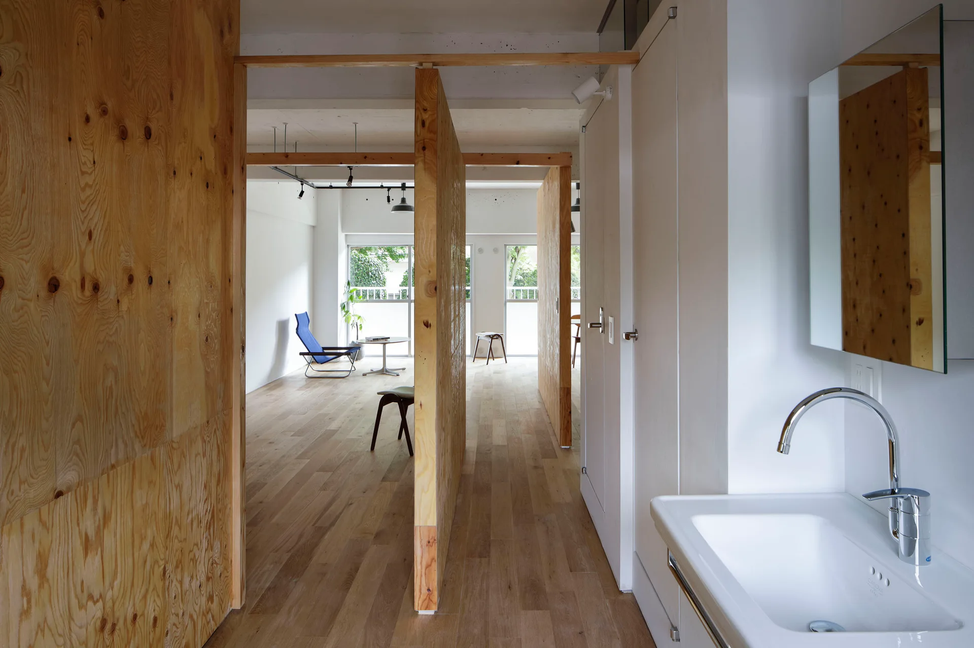 An open apartment interior features raw plywood partitions, light wood floors, a white bathroom sink, and a distant blue lounge chair.
