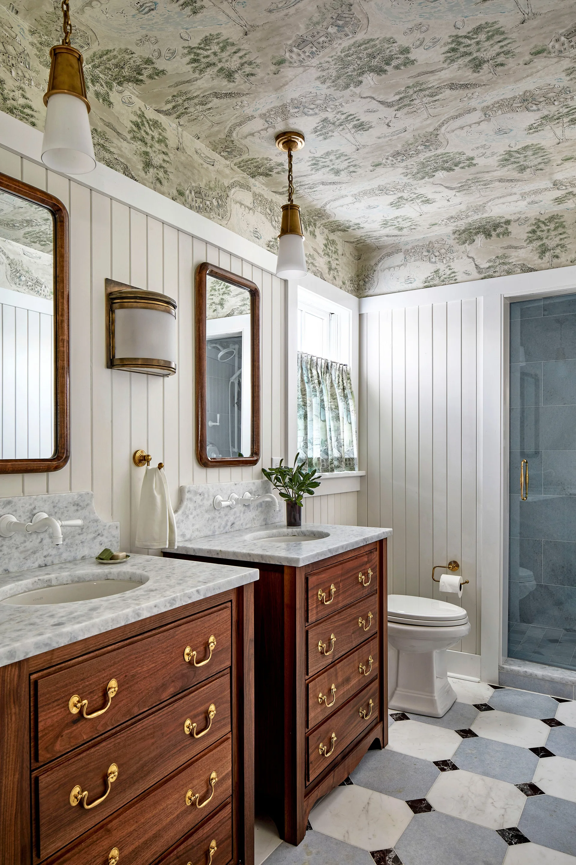 A bathroom features dual wood vanities with marble tops, vertical white wall paneling, and a green scenic wallpaper covering the ceiling.