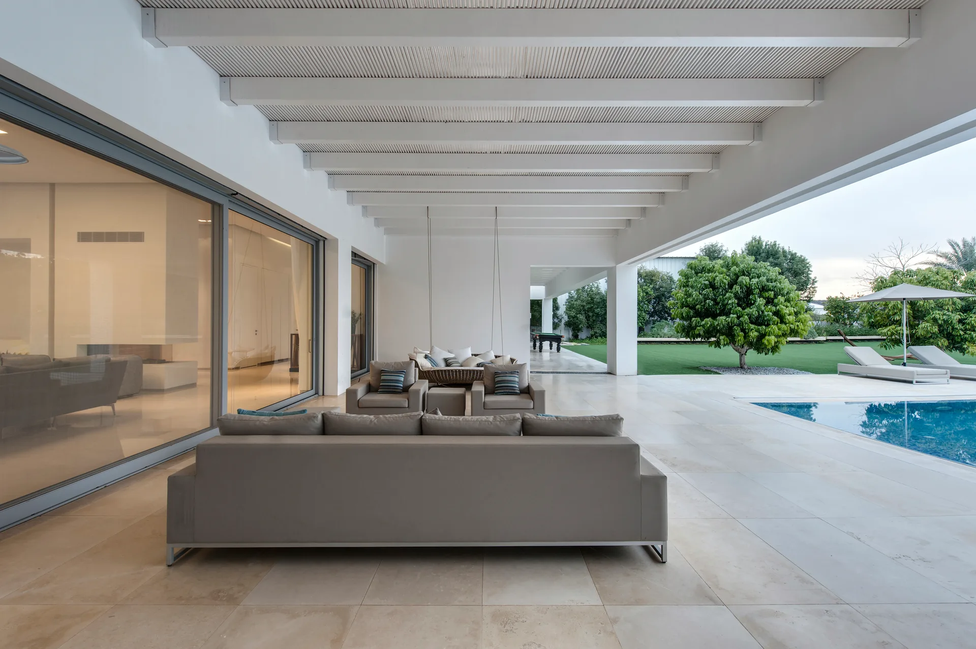 A modern covered patio features grey outdoor sofas, white exposed ceiling beams, and large format stone tiles next to a swimming pool.