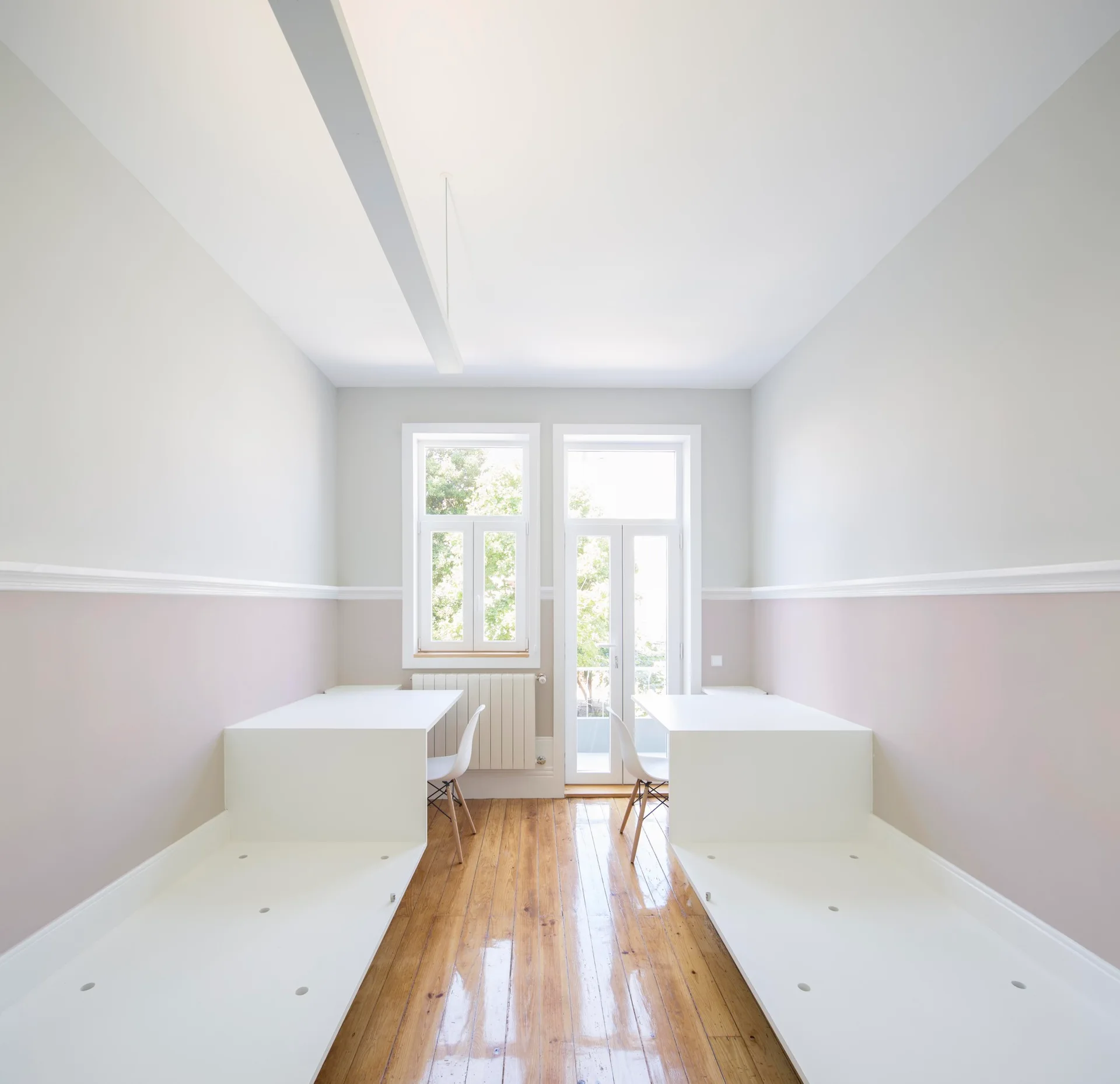 A symmetrical room features two built-in white platform desks with white chairs on a glossy wood floor under a two-tone mauve and white wall.