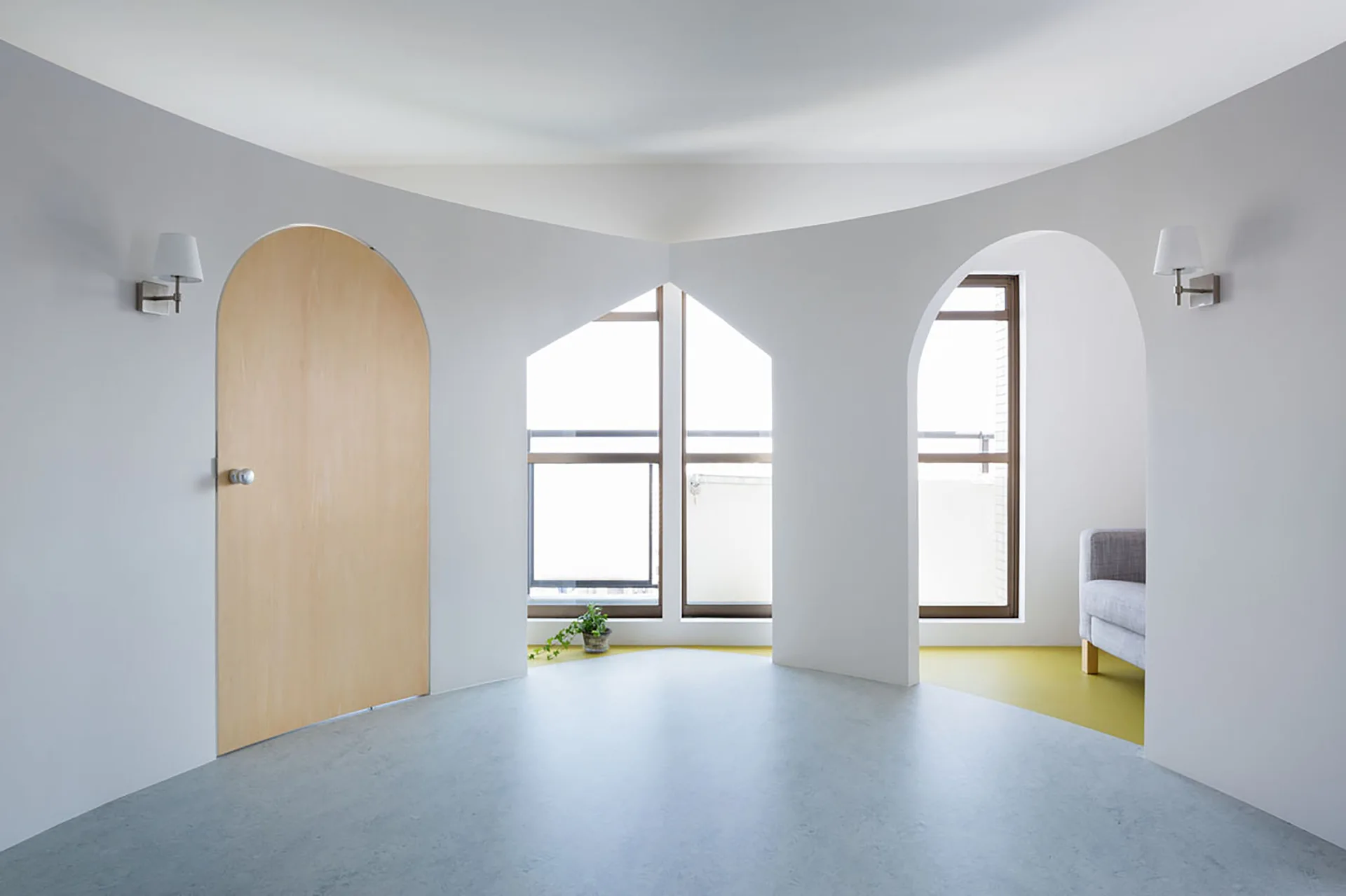 A modern minimalist room featuring curved white walls with arched doorways, a light wood arched door, and a split grey and yellow floor.