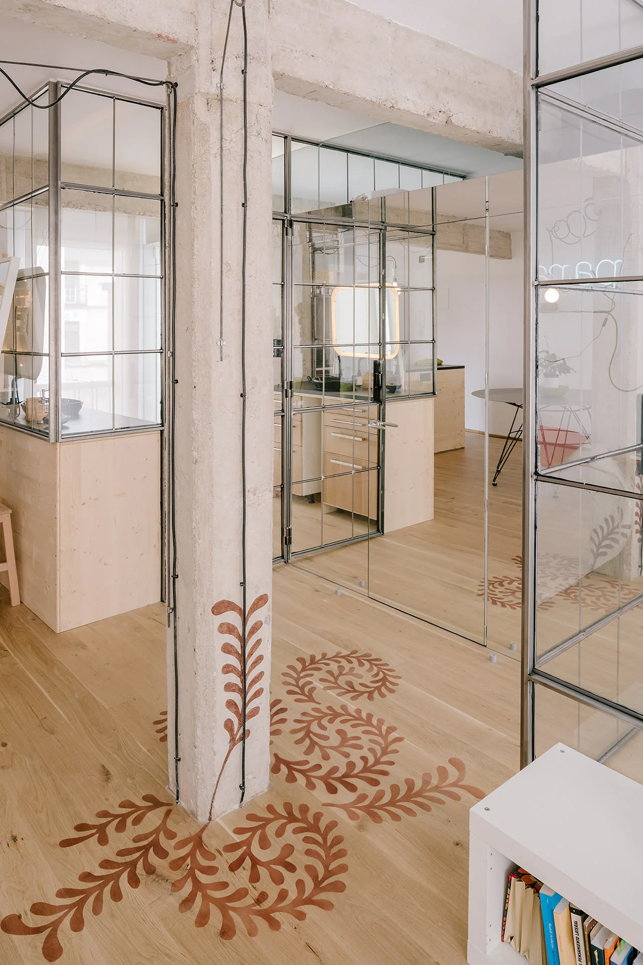 An industrial interior features steel-framed glass partitions, light wood floors, and an exposed concrete column decorated with a terracotta botanical motif.