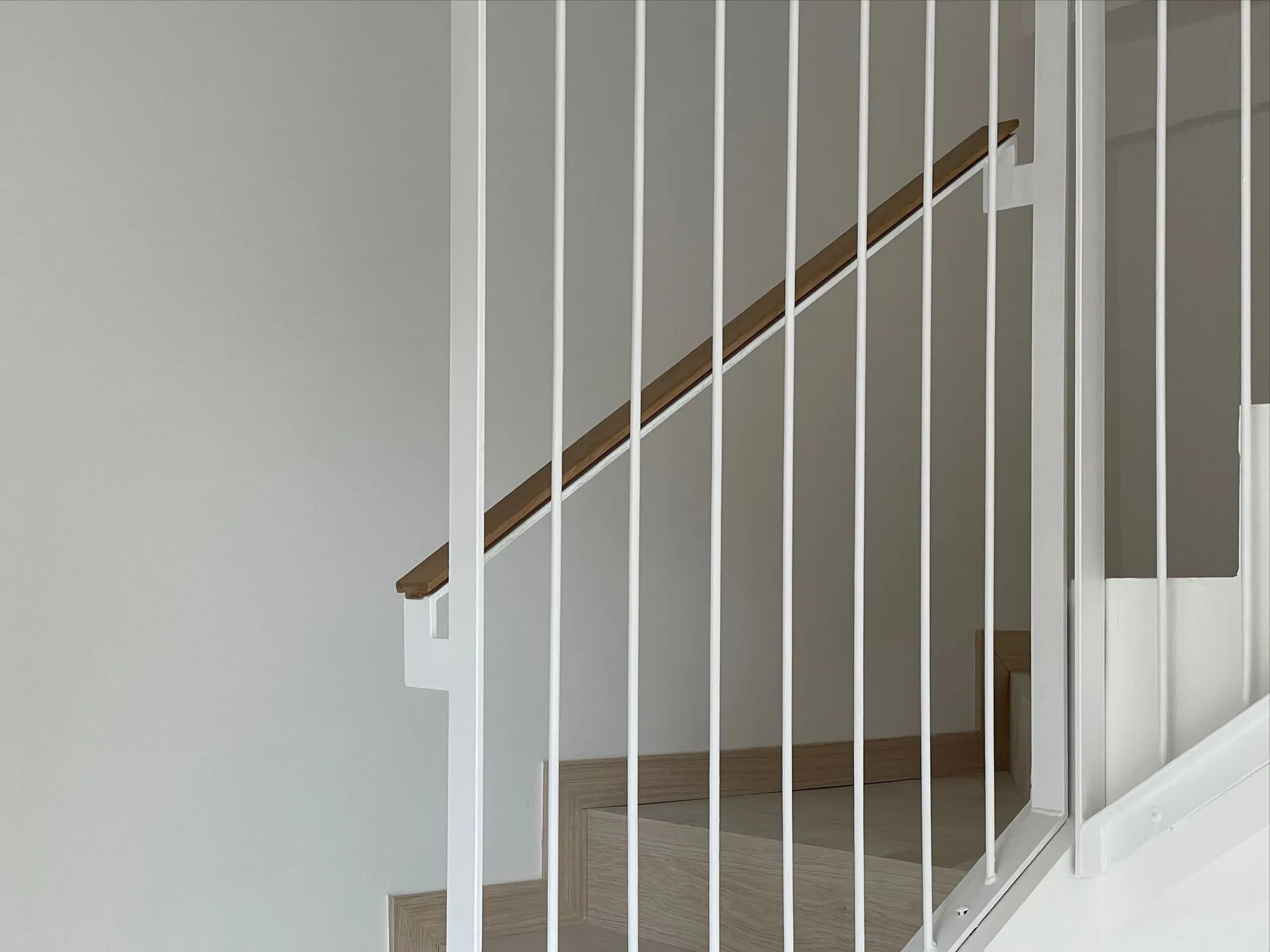 A modern staircase features vertical white metal balusters, a natural wood handrail, and matching wood steps against a neutral wall.