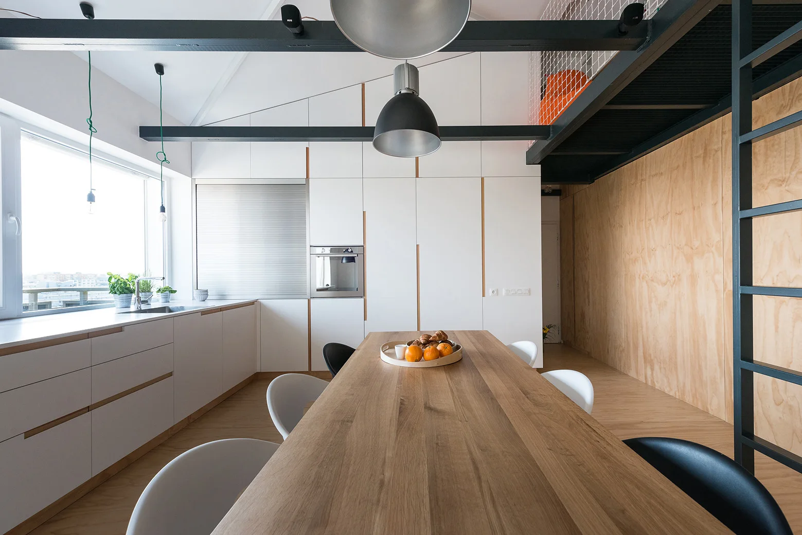 A minimalist kitchen and dining space with a long wooden table, white cabinets, and structural black ceiling beams.