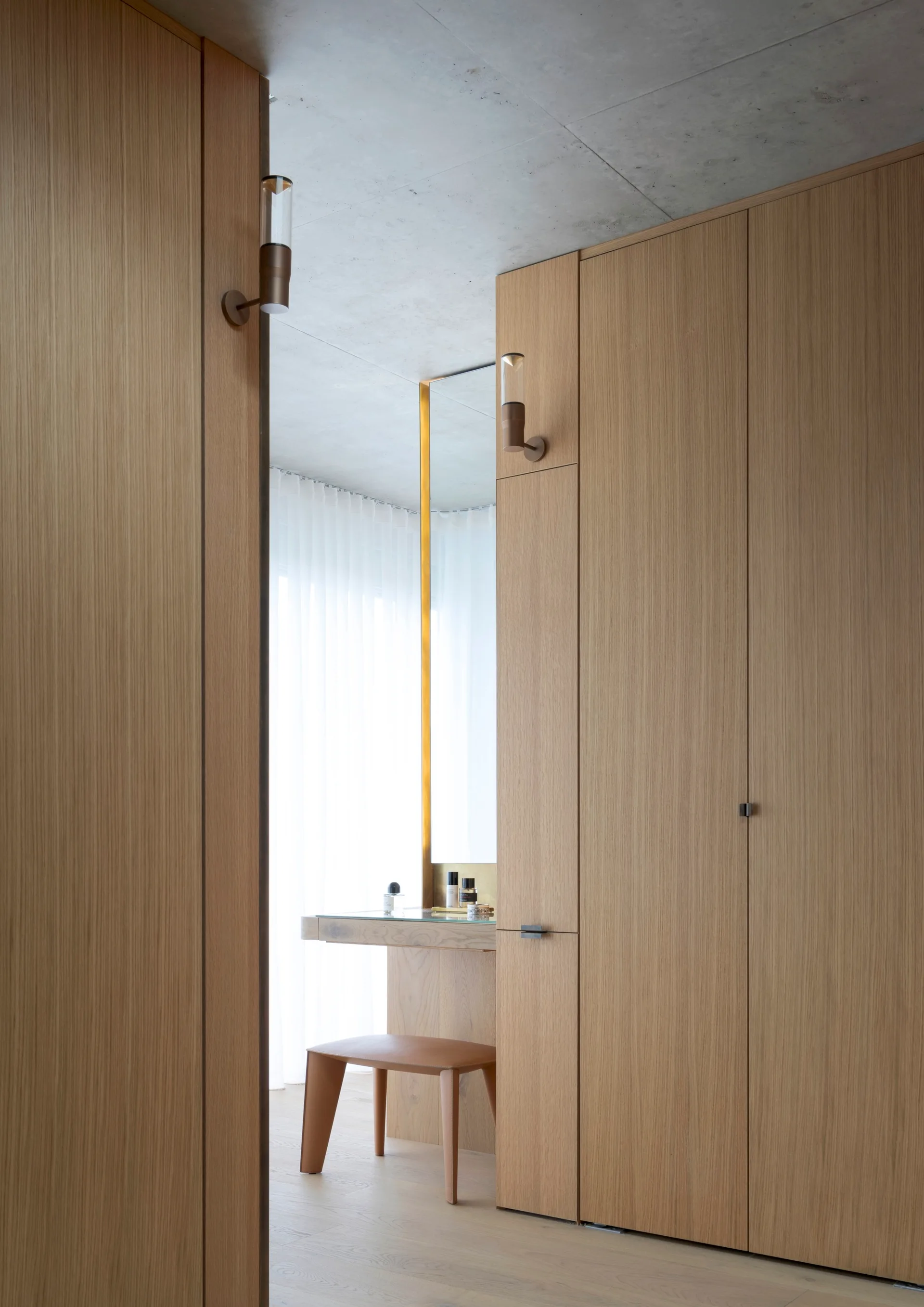 A walk-in dressing room features full-height wood closets, a built-in vanity table with a leather stool, and two brass sconces below a concrete ceiling.